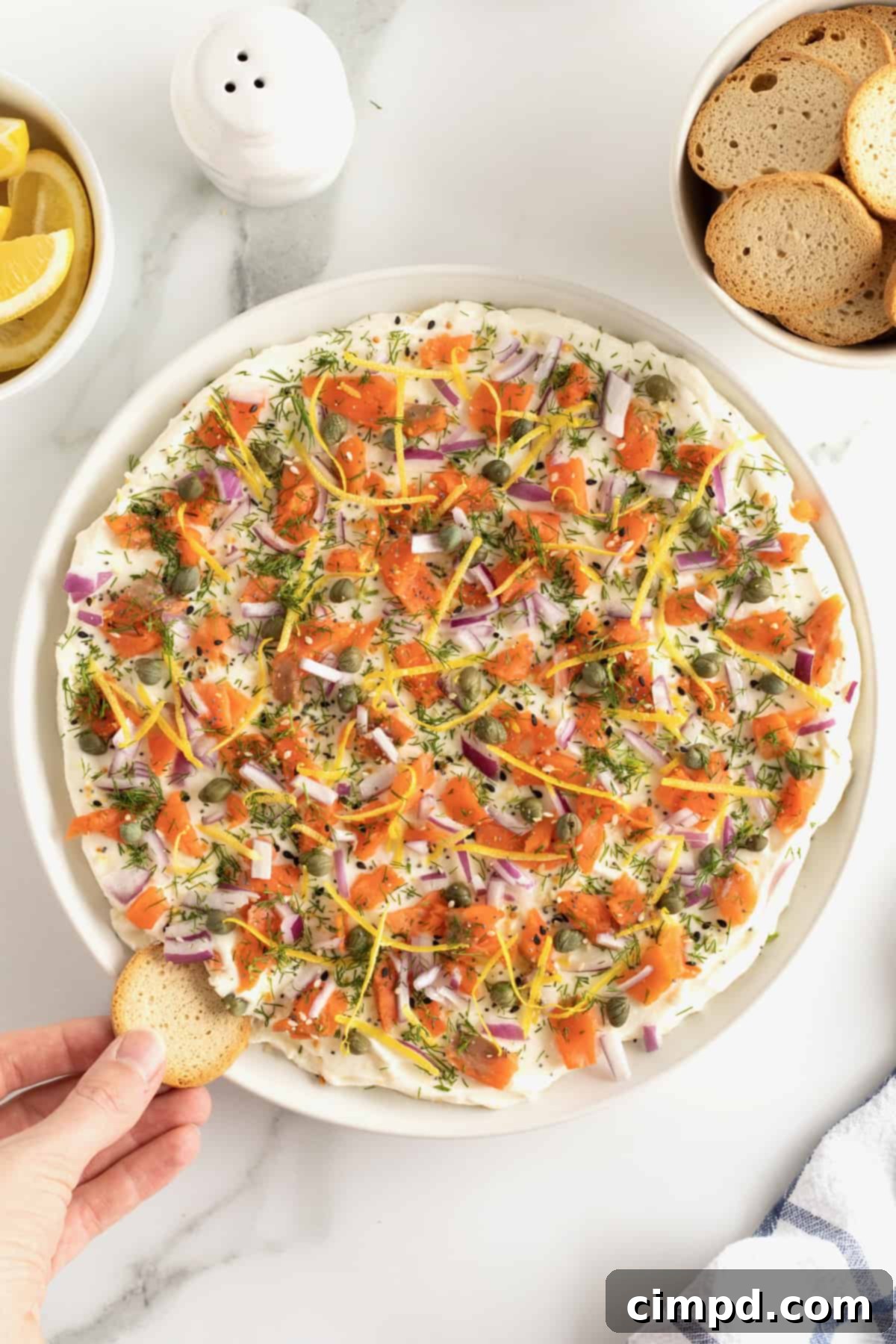 A round white serving platter with cream cheese smoked salmon dip, garnished with red onion, capers, and lemon zest, with bagel chips on the side.