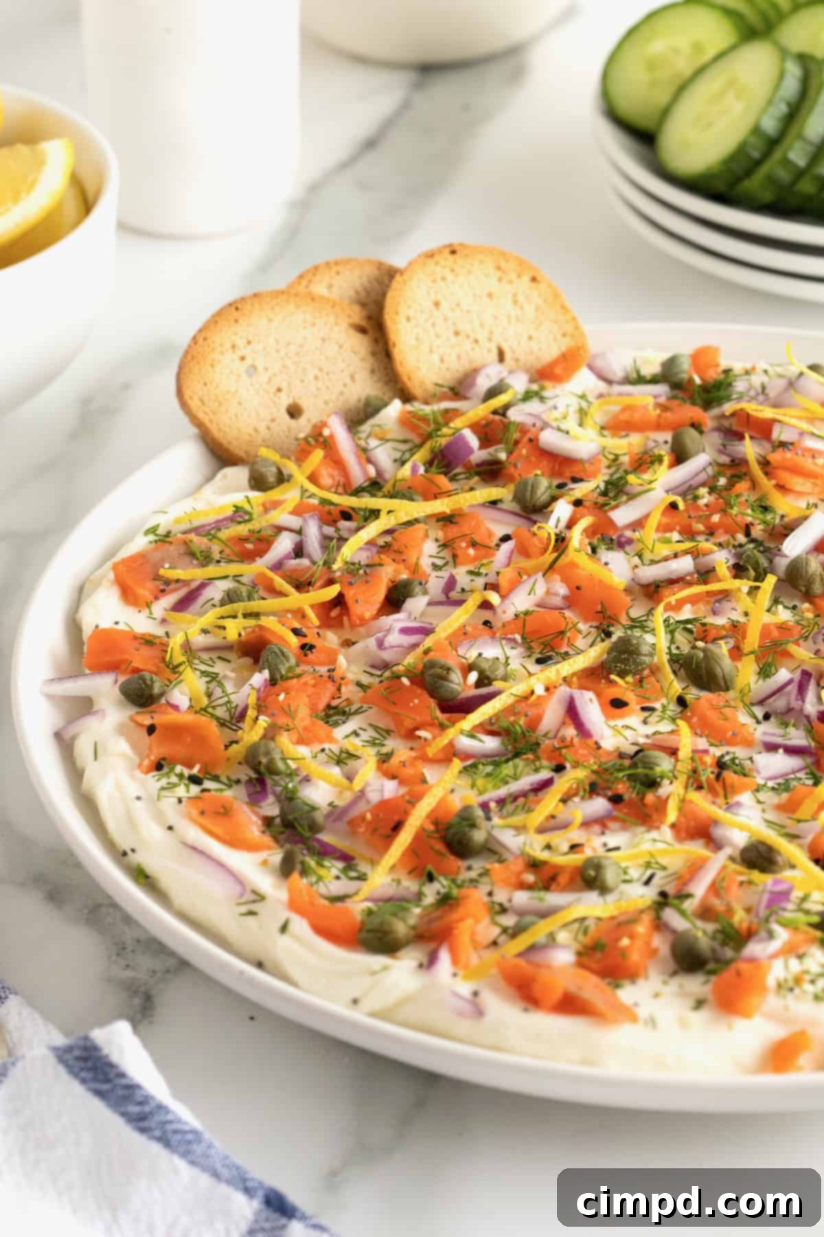 A vibrant close-up of the Smoked Salmon Bagel Dip, highlighting the fresh dill, capers, and lemon zest atop the creamy base, surrounded by bagel chips.