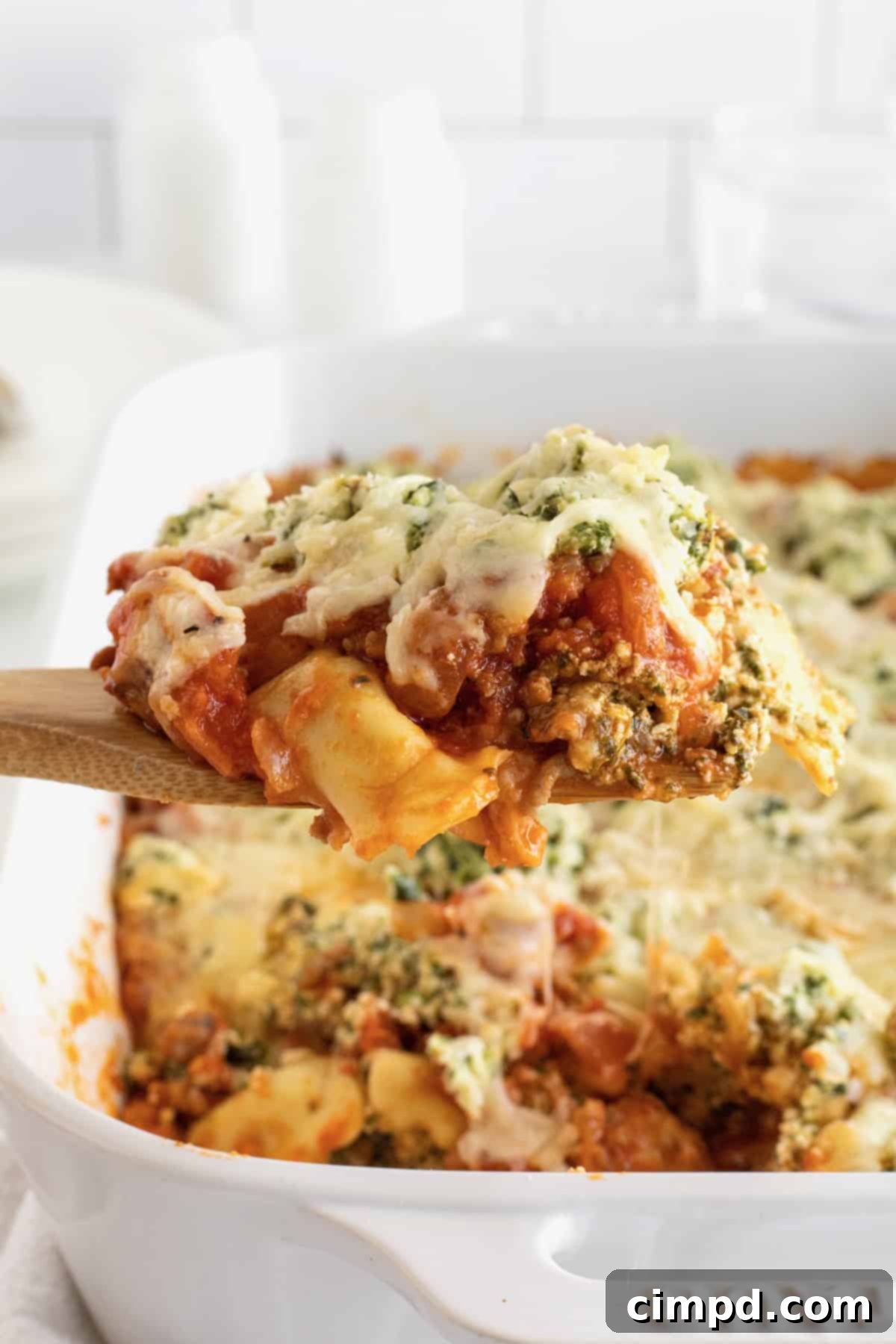A wooden spoon holding pasta covered in tomato sauce, melted mozzarella cheese and fresh herbs over a white casserole dish of lasagna.