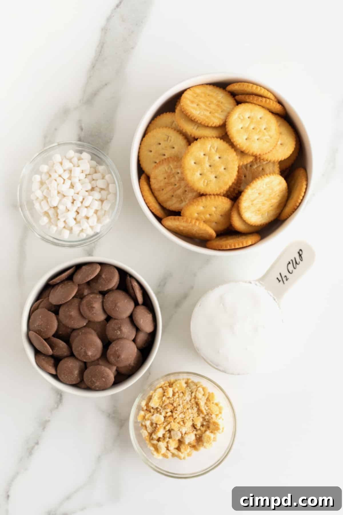 No-Bake Smores Cracker Bites 5 A flat lay photograph displaying all the ingredients for S'mores Cracker Bites: Ritz crackers, marshmallow bits, crushed cracker crumbs, marshmallow creme, and chocolate candy wafers, neatly arranged in small glass dishes on a white marble countertop.