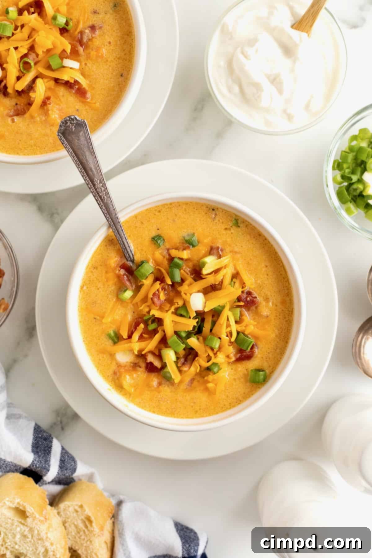A beautifully presented white bowl of baked sweet potato soup, garnished with crispy bacon bits, melted shredded cheddar cheese, and vibrant sliced green onions, resting on a white saucer with a spoon. A true comfort food delight.