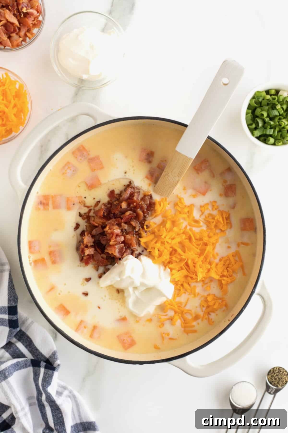 A white Dutch oven filled with creamy sweet potato soup, with bacon bits, a dollop of sour cream, and shredded cheddar cheese visible before being stirred in, accompanied by a wooden spoon.