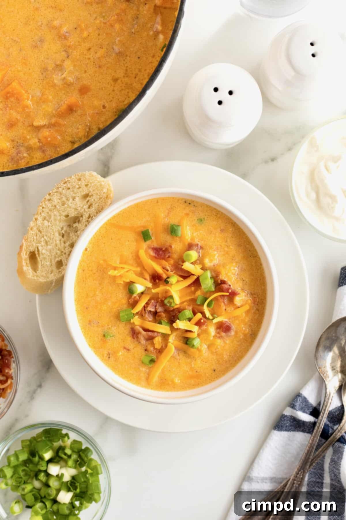 A comforting white bowl of sweet potato soup, topped with crispy bacon, shredded cheese, and green onions, served with a spoon and a slice of French bread on a white saucer, illustrating a perfect leftover meal.