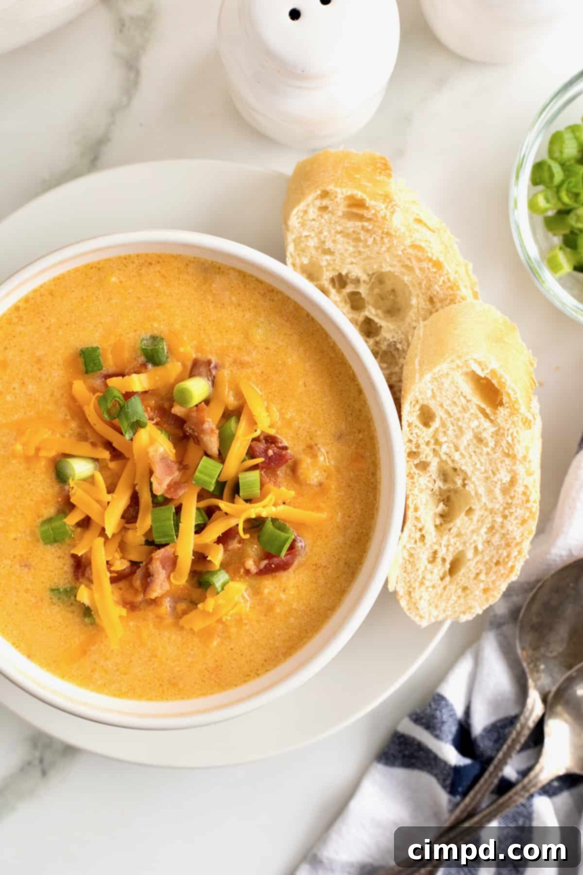 A white bowl of baked sweet potato soup, generously topped with crispy bacon, shredded yellow cheddar cheese, and fresh green onions, sits on a white saucer alongside two slices of toasted French bread, ready for a comforting meal.