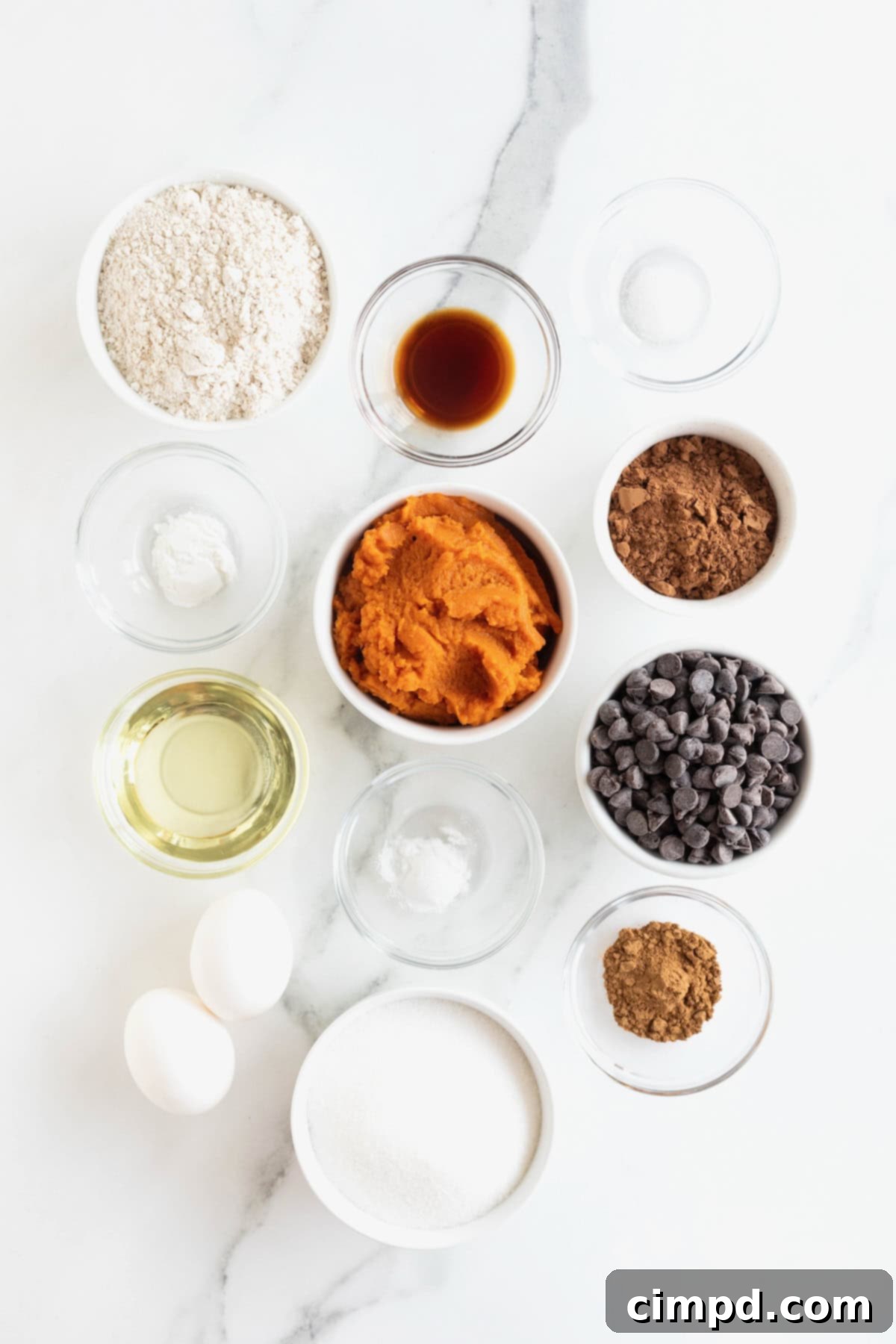 Assorted ingredients for whole wheat pumpkin brownies, neatly arranged in small glass containers on a white marble counter, ready for mixing.