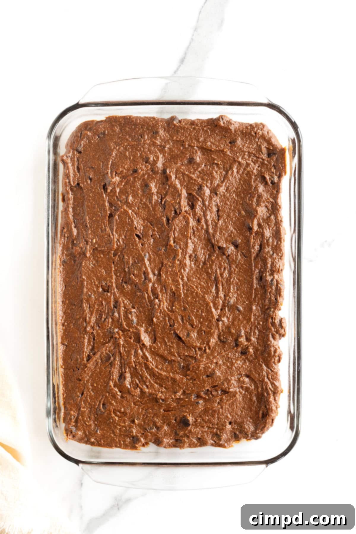 Smooth pumpkin brownie batter evenly spread in a glass baking dish on a white marble counter, ready for the oven.
