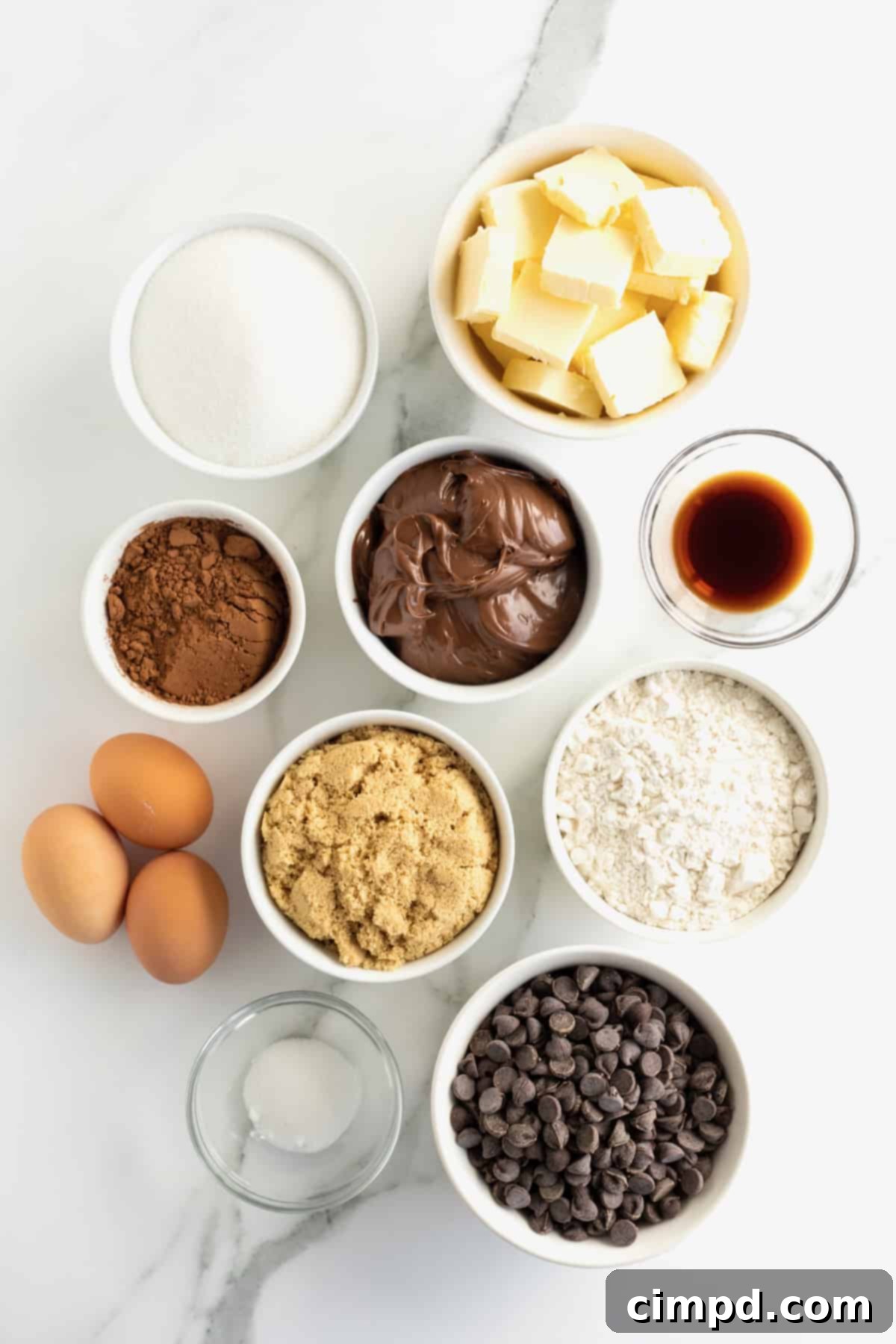 Butter, chocolate chips, granulated sugar, brown sugar, cocoa powder, vanilla, All-purpose flour, kosher salt, Nutella chocolate hazelnut spread in small containers on a white counter top next to three large brown eggs.