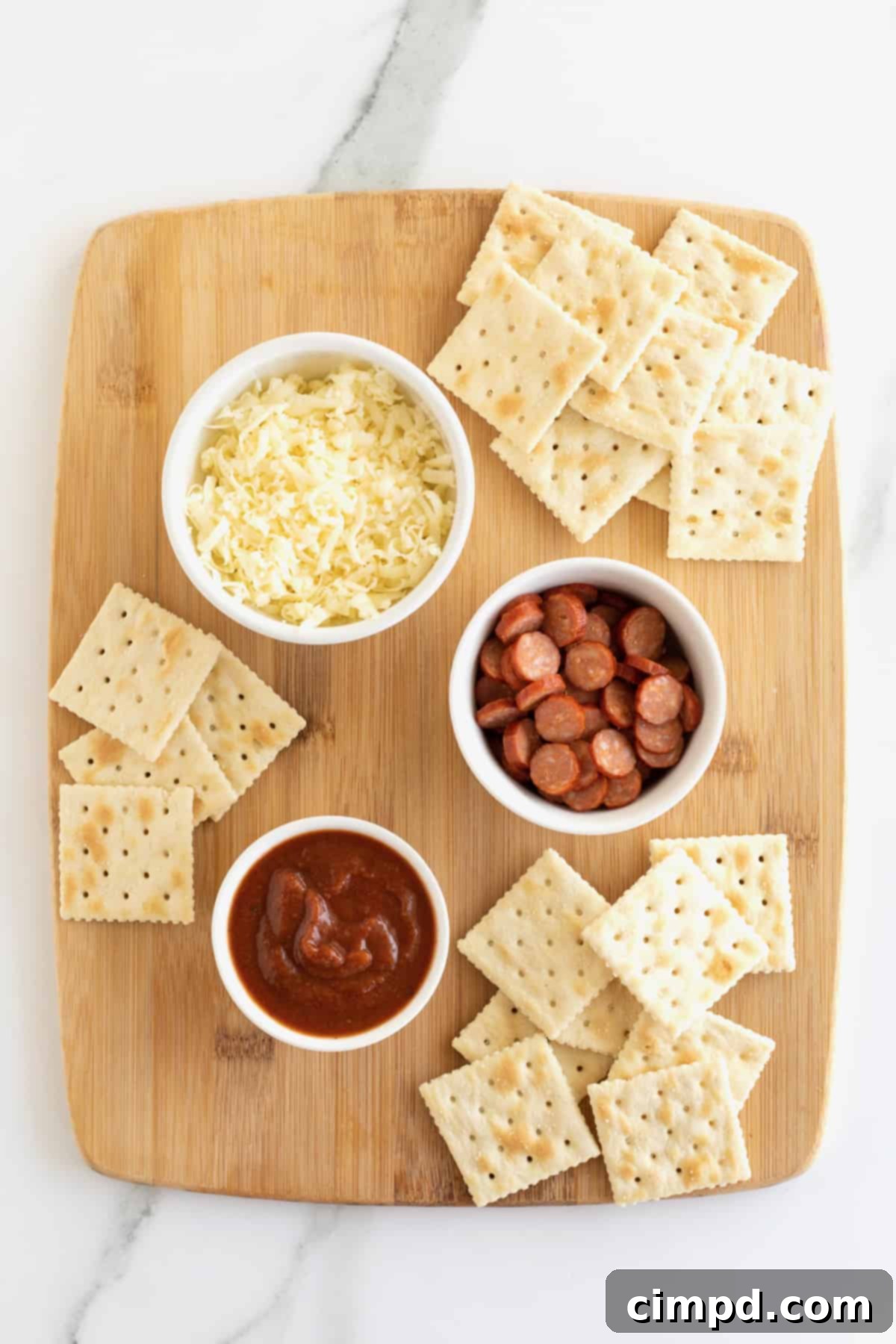Crispy Pizza Bites 5 Ingredients for pizza cracker bites: shredded mozzarella cheese, mini pepperoni, pizza sauce, and saltine crackers, arranged on a wooden cutting board.