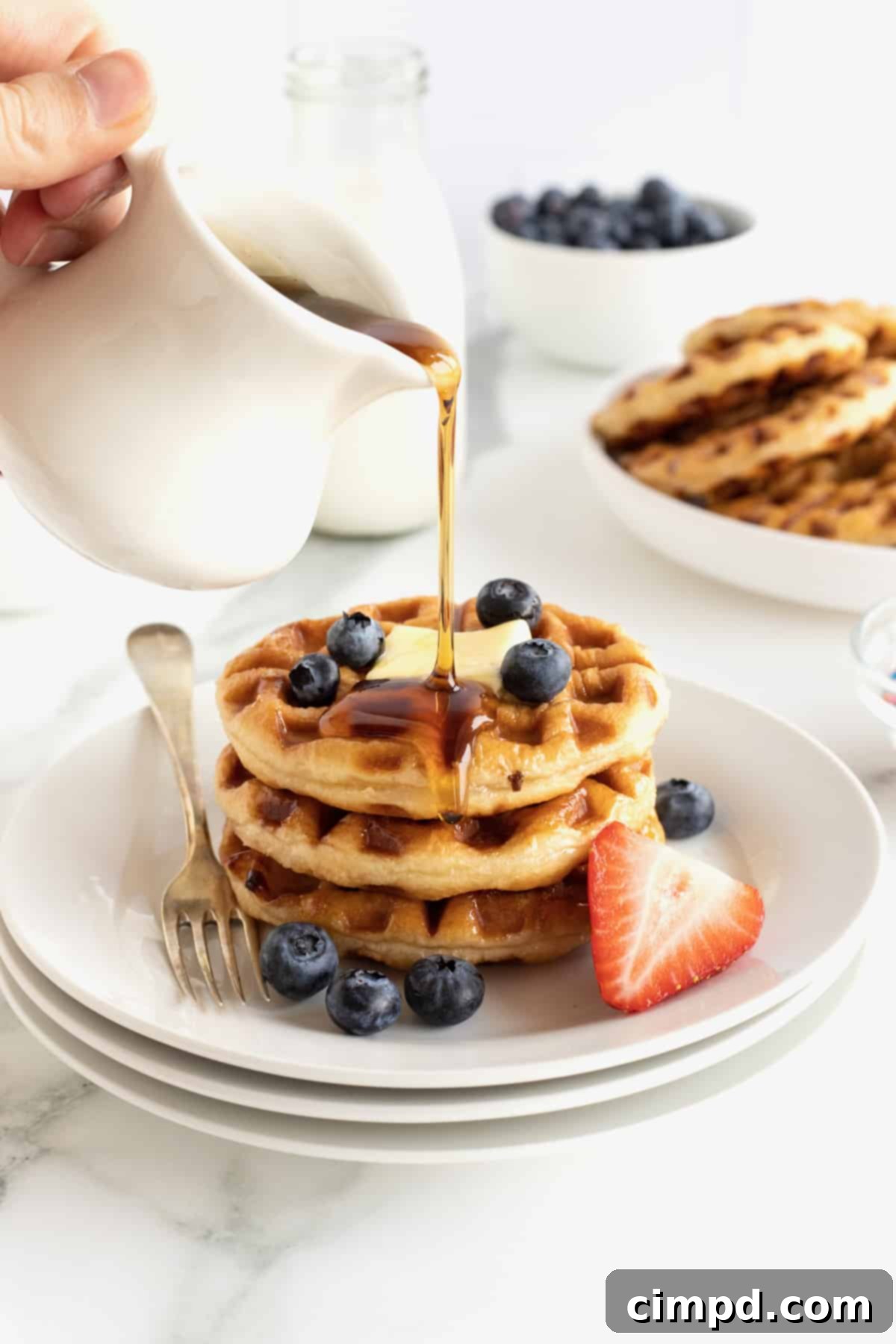 Glazed Waffle Rings 3 Three donut waffles stacked on a white plate with a square of butter, fresh blueberries and hot maple syrup poured over the top.