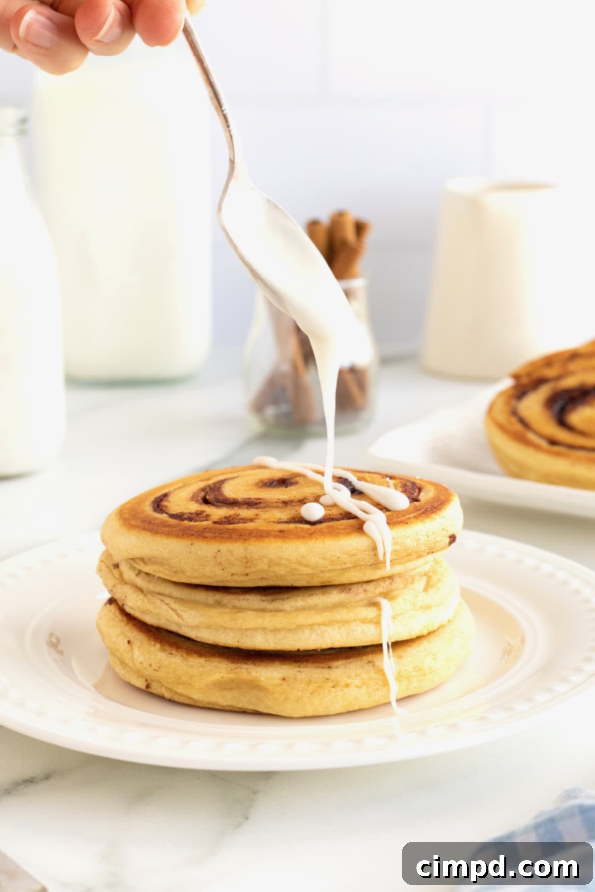 Cinnamon Swirl Pancakes 3 Icing being drizzles onto a stack of three cinnamon roll pancakes on a white plate.