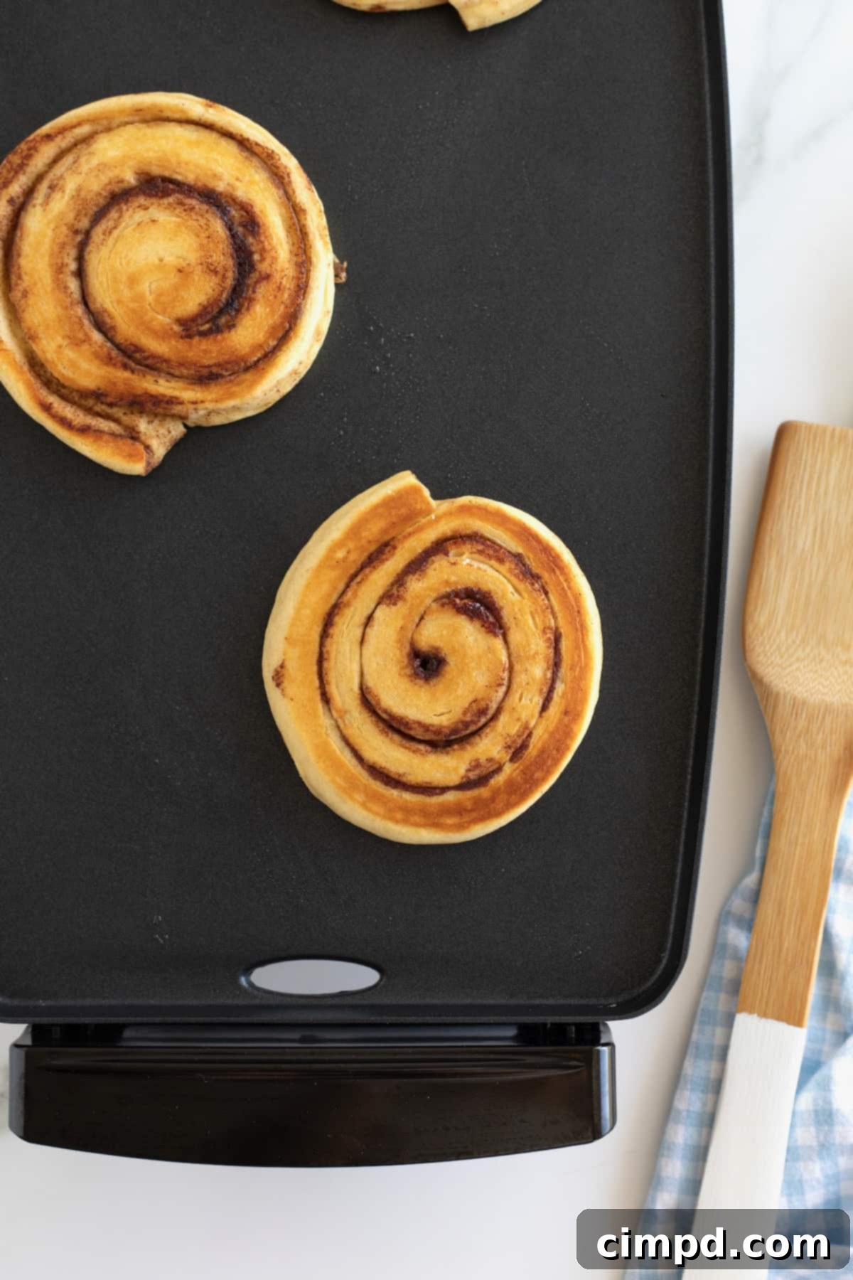 Cinnamon Swirl Pancakes 4 Two cinnamon roll pancakes on a nonstick griddle.