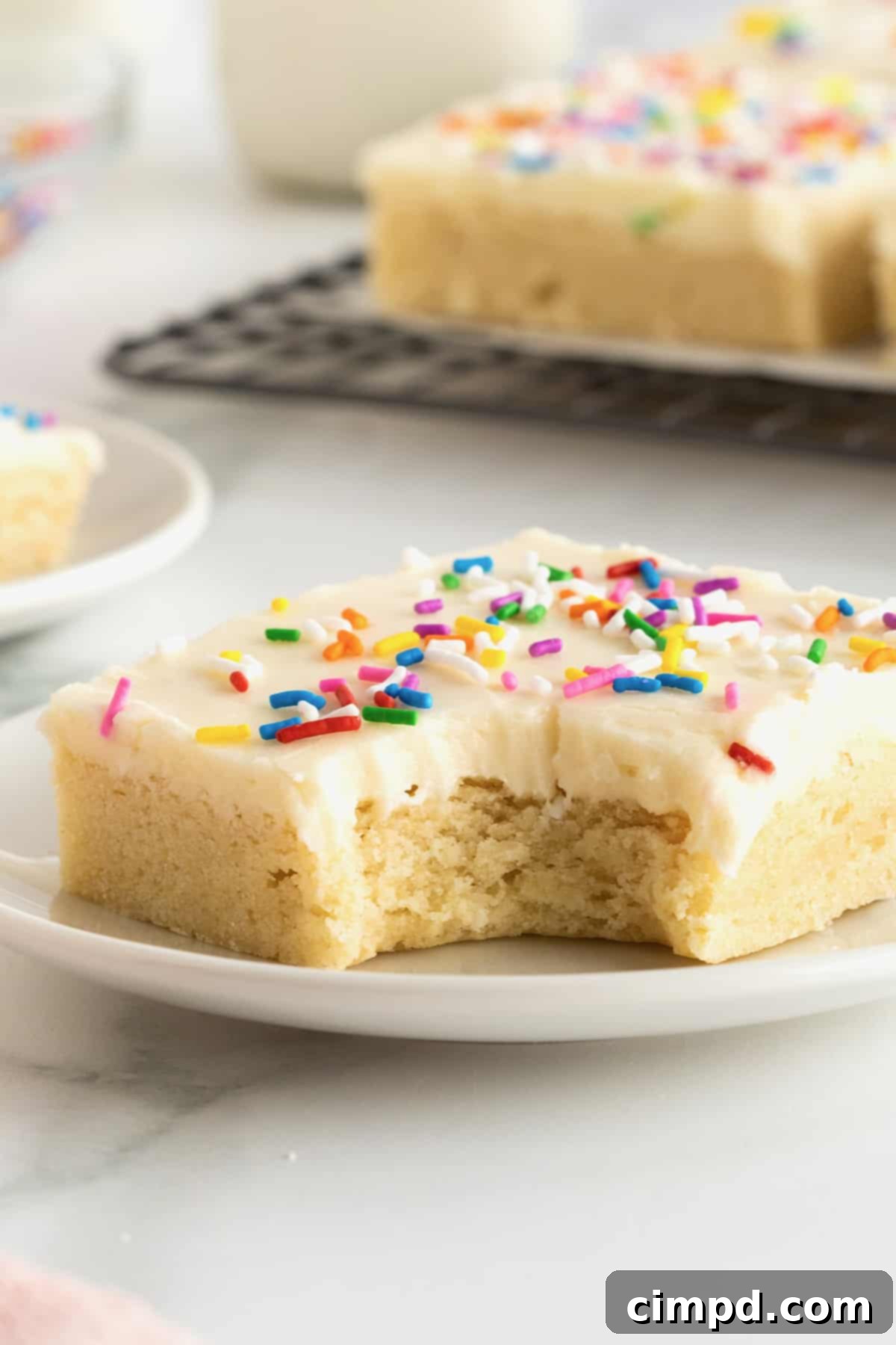Soft Frosted Sugar Cookie Bars 3 A square sugar cookie with frosting and rainbow sprinkles on a white plate.