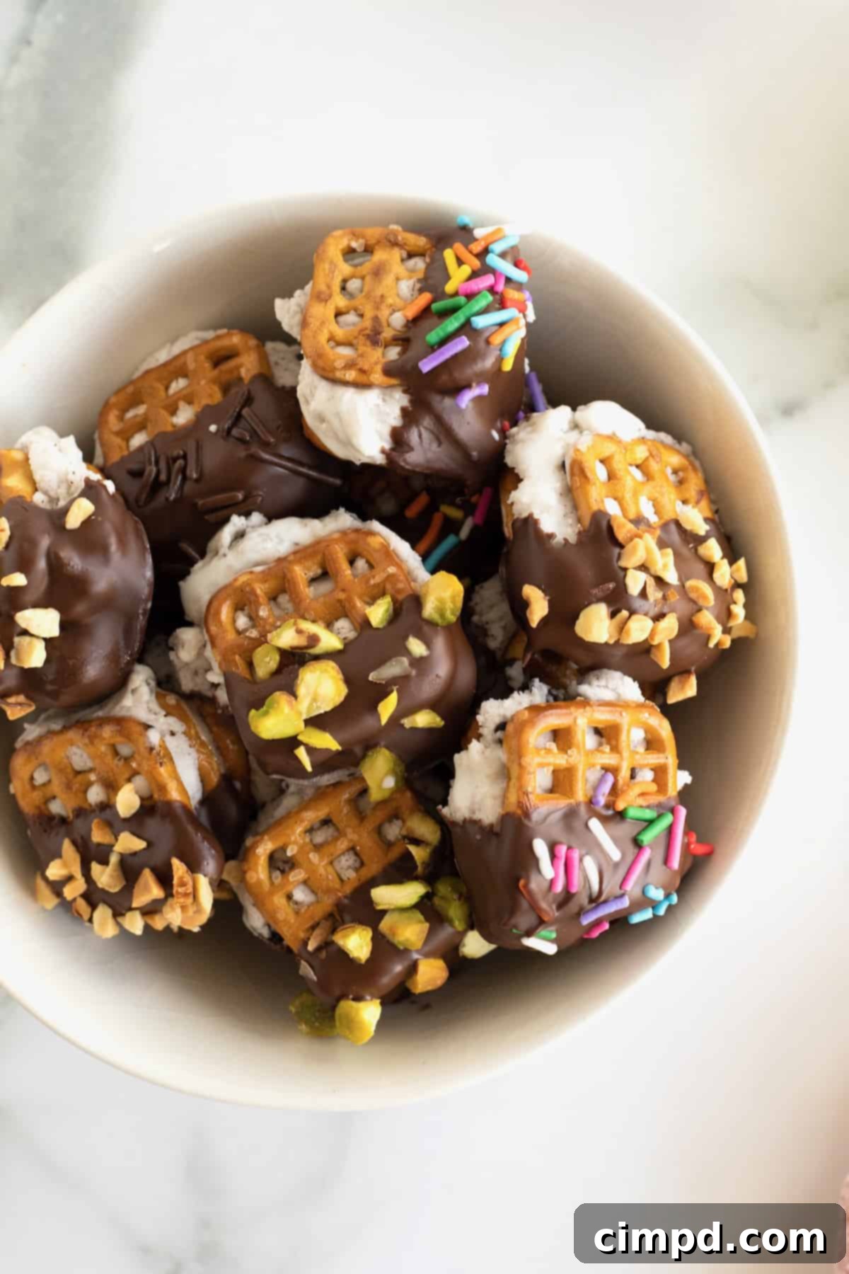 A white serving bowl filled with pretzel ice cream sandwich bites dipped in chocolate and coated in sprinkles.