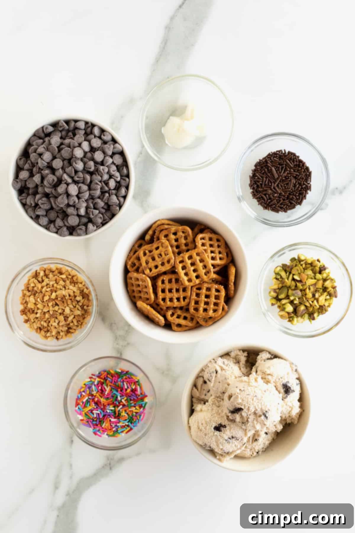 Pretzel snaps, cookies and cream ice cream, chocolate chips, rainbow sprinkles, chopped peanuts, and chopped pistachios in small glass bowls.