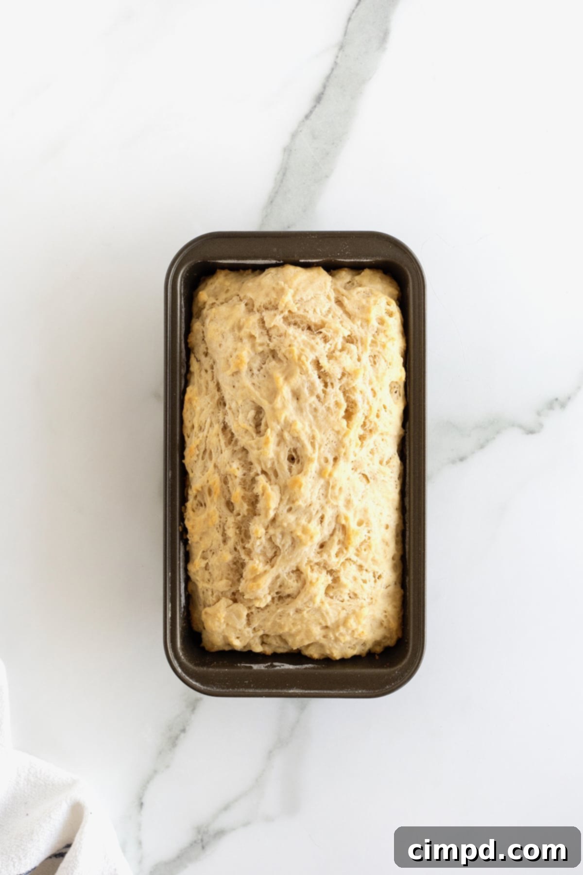 Pub-Style Beer Bread 11 A freshly baked loaf of golden-brown beer bread, still in its dark metal loaf pan, cooling on a white marble counter.