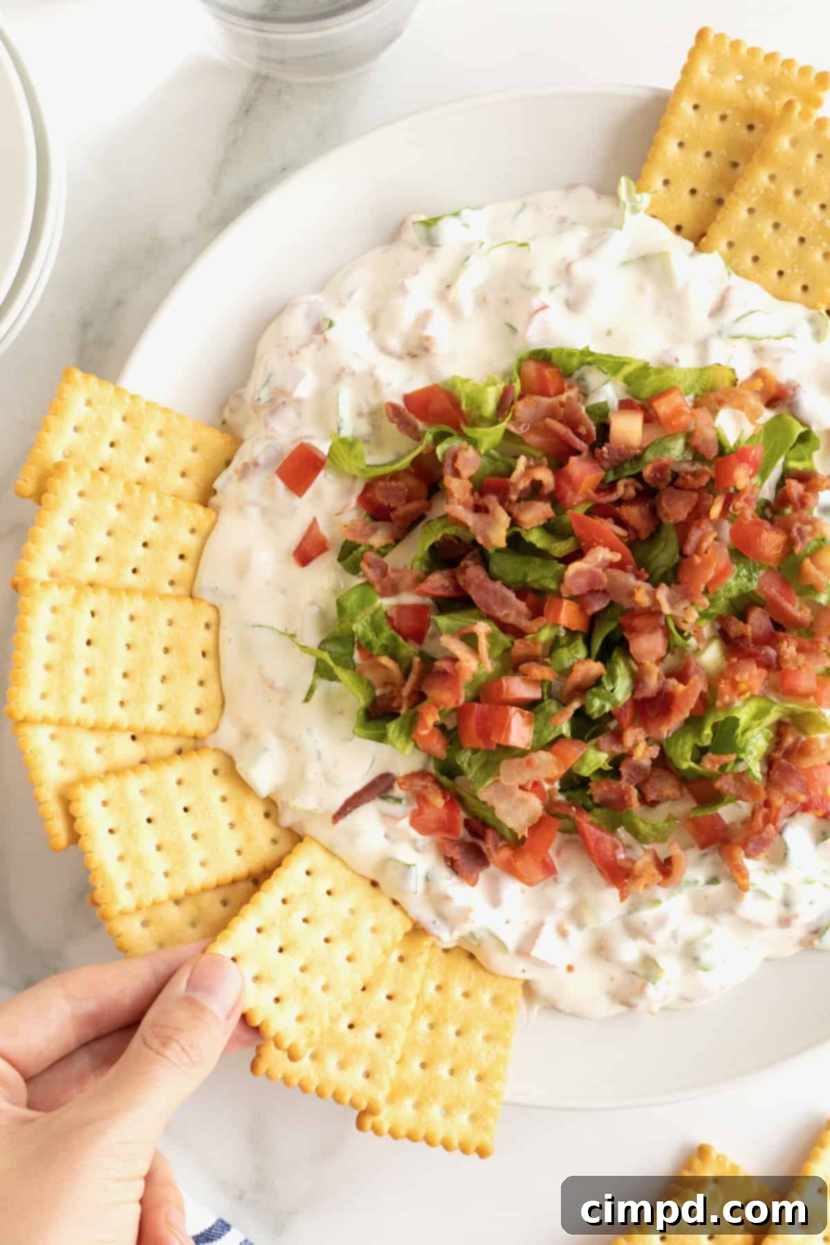 Ultimate BLT Dip 2 A beautifully presented white serving bowl filled with creamy BLT Dip, garnished generously with crumbled bacon and shredded lettuce, accompanied by a side of buttery club crackers.