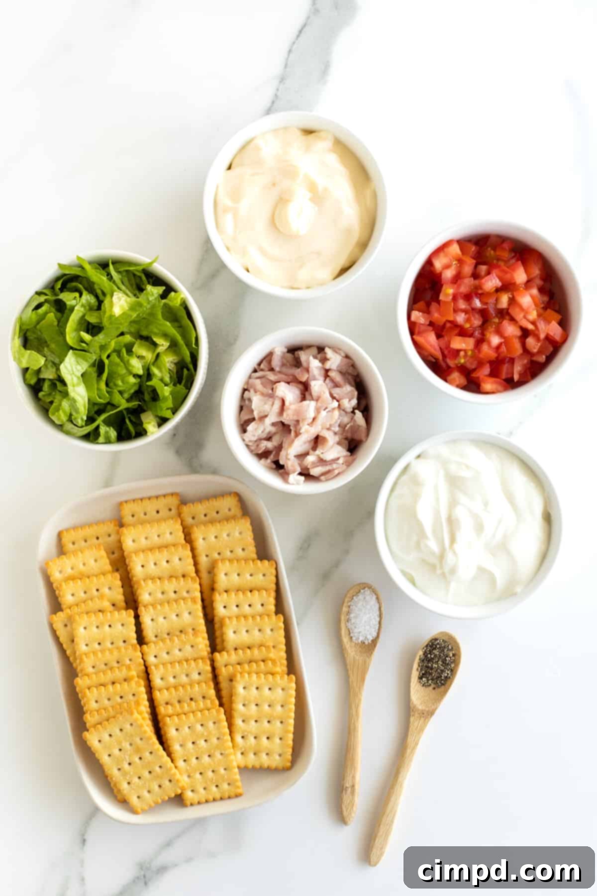 Ultimate BLT Dip 6 A flat lay photograph displaying all the fresh ingredients for BLT dip: crispy cooked bacon, vibrant romaine lettuce, creamy sour cream, juicy Roma tomatoes, rich mayonnaise, and a stack of club crackers.