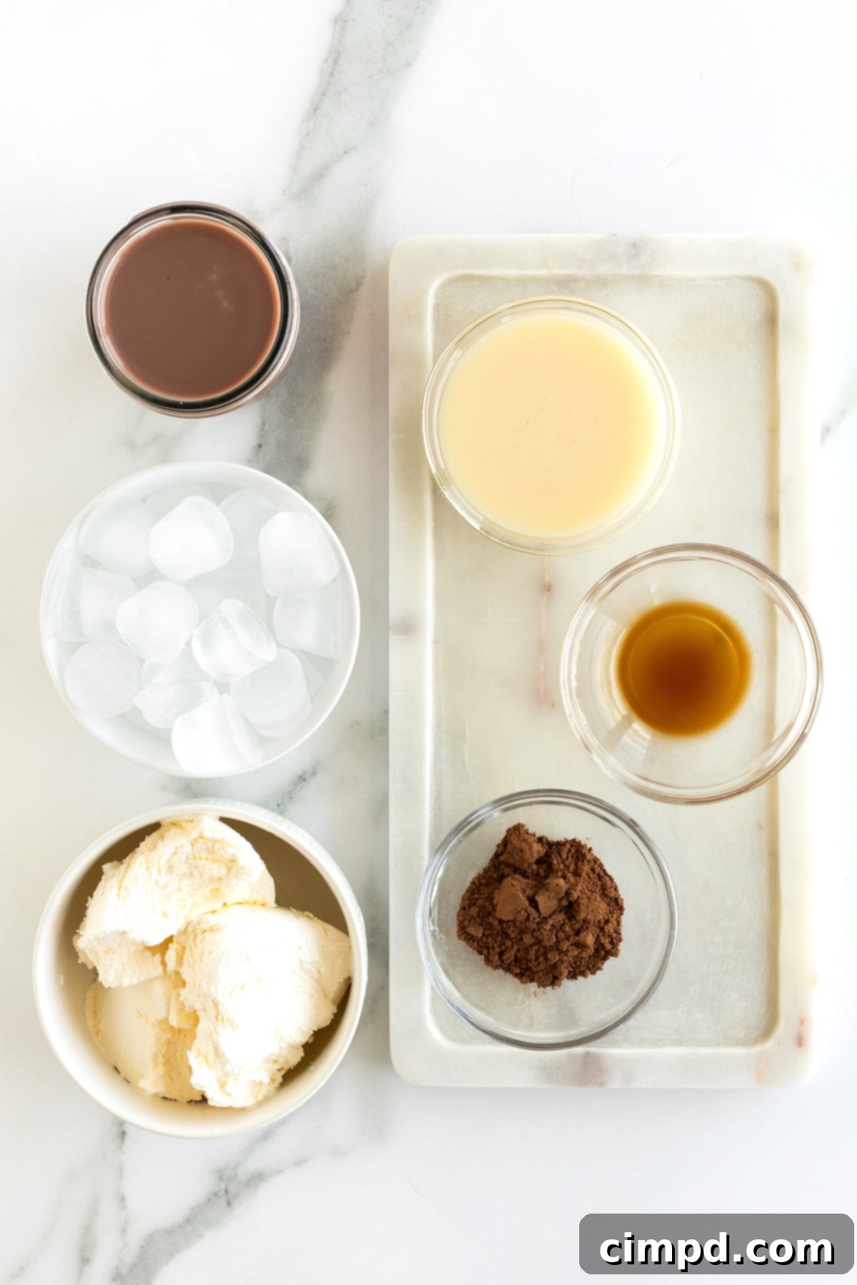 Cocoa powder, vanilla extract, sweetened condensed milk, Cool Whip, chocolate milk, and ice neatly arranged in glass containers on a kitchen counter, ready for blending into a homemade Wendy's Frosty.
