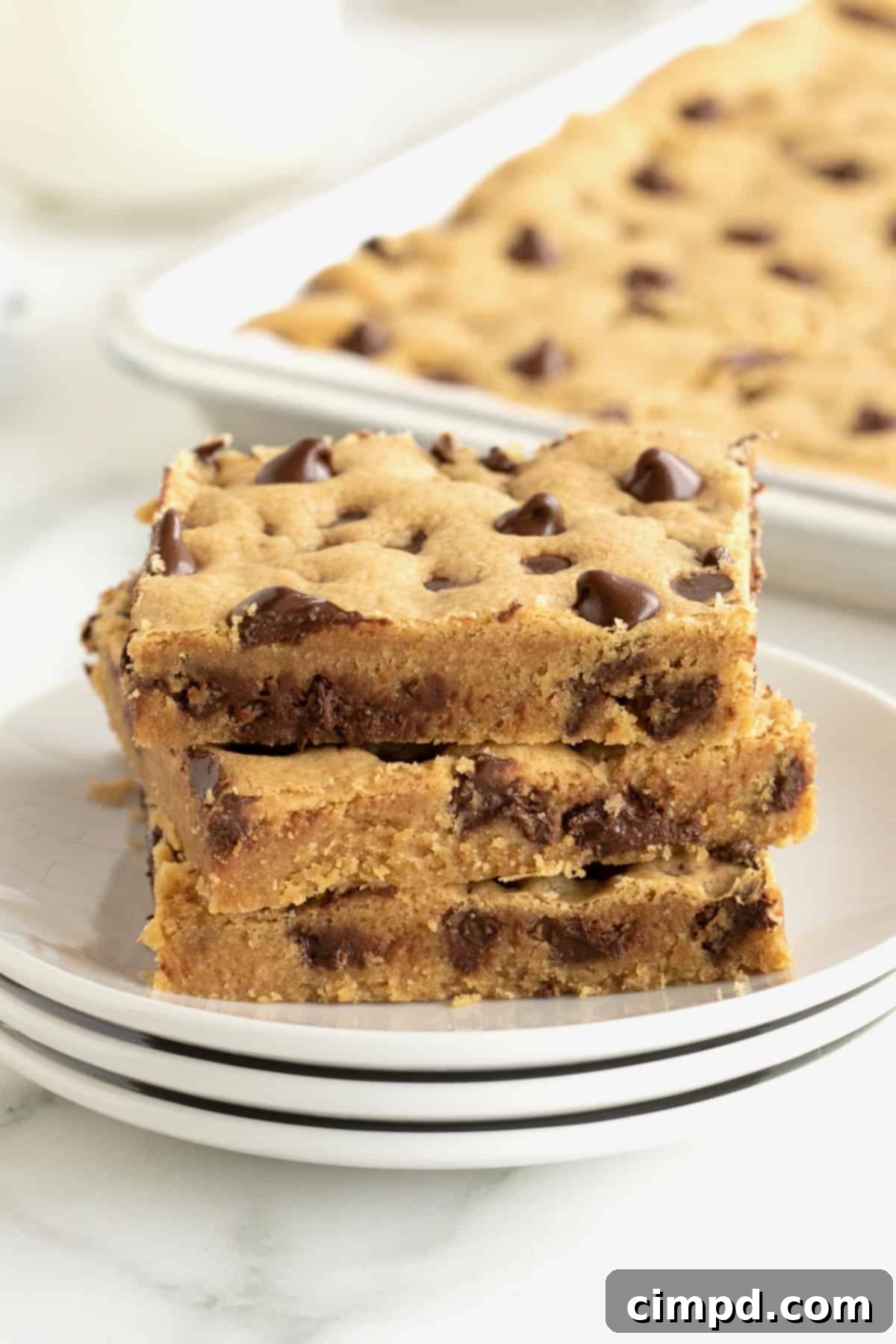 A stack of three peanut butter cookie bars with chocolate chips on a stack of three white plates with a silver sheet pan of cookie bars partially visible in the background.