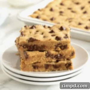 A stack of three peanut butter cookie bars with chocolate chips on a stack of three white plates with a silver sheet pan of cookie bars partially visible in the background.