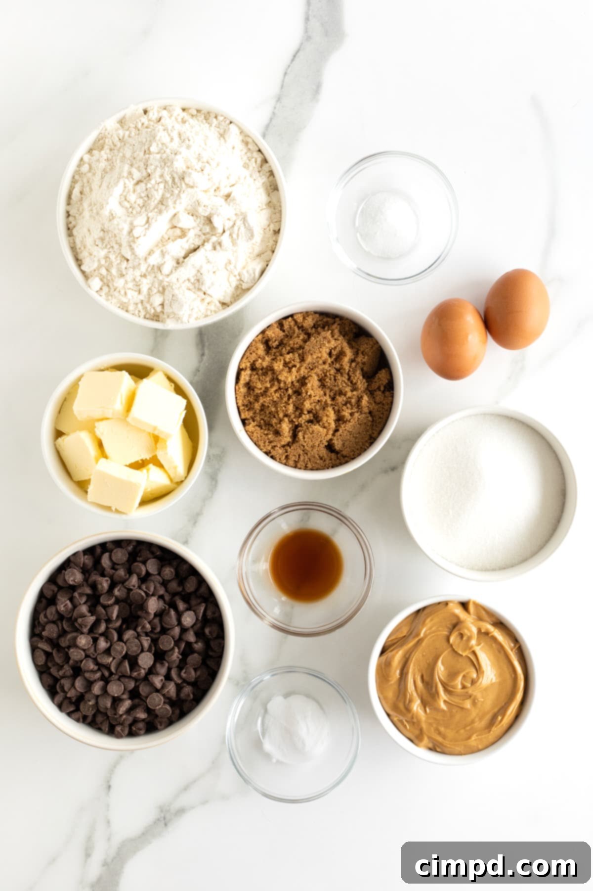 Butter, sugar, flour, peanut butter, dark brown sugar, vanilla extract and chocolate chips in small glass containers on a counter next to two brown eggs.