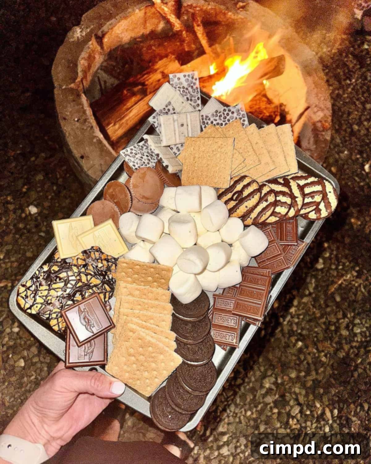 A baking tray filled with a variety of s'mores ingredients including marshmallows, graham crackers, chocolate bars, and fudge striped cookies, set in front of a warm fire pit.