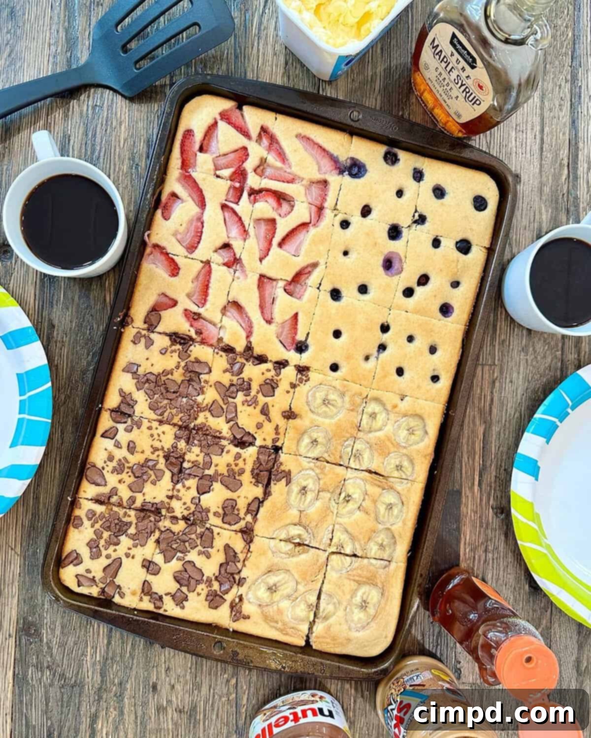 A baking sheet filled with golden, fluffy sheet pan pancakes, ready to be served, accompanied by two steaming cups of coffee on a wooden surface.