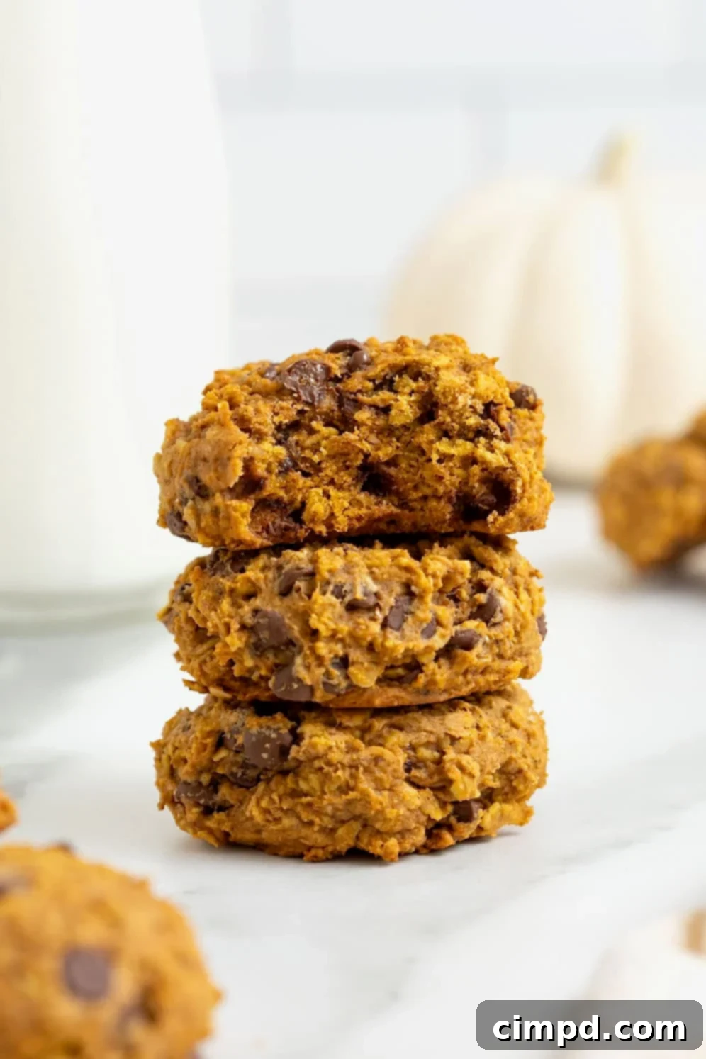 A stack of three pumpkin oatmeal chocolate chip cookies on a white marble counter. The top cookie has a bite out of it.