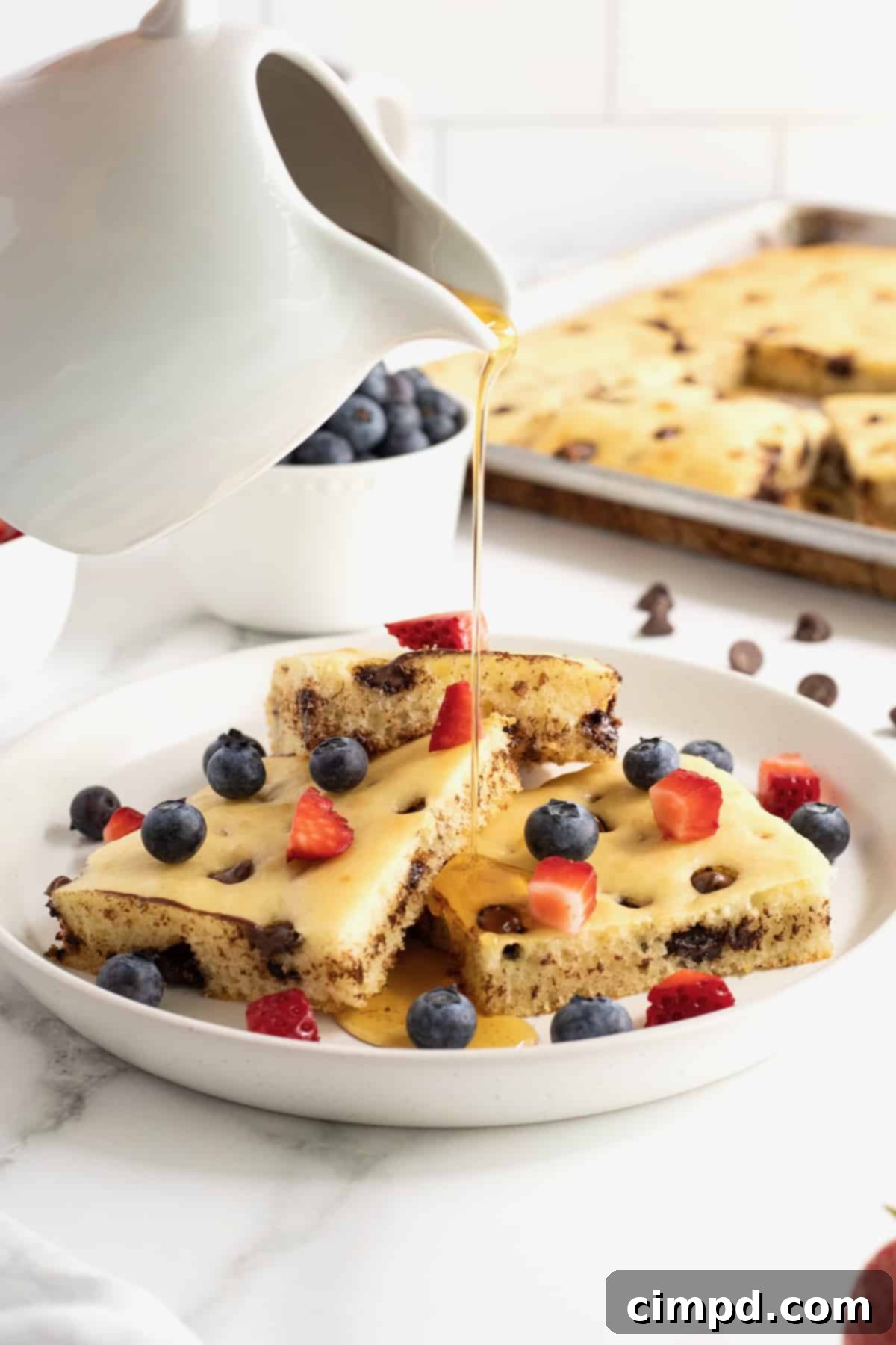 Syrup being poured from a white pitcher over a plate of square-cut sheet pan chocolate chip pancakes, garnished with vibrant diced strawberries and blueberries.