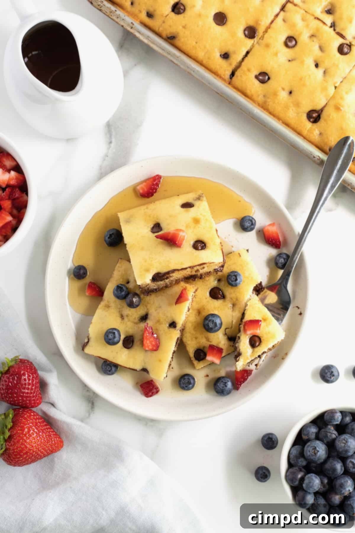 A beautifully arranged plate of square-cut sheet pan chocolate chip pancakes, generously drizzled with maple syrup and topped with colorful diced strawberries and blueberries.