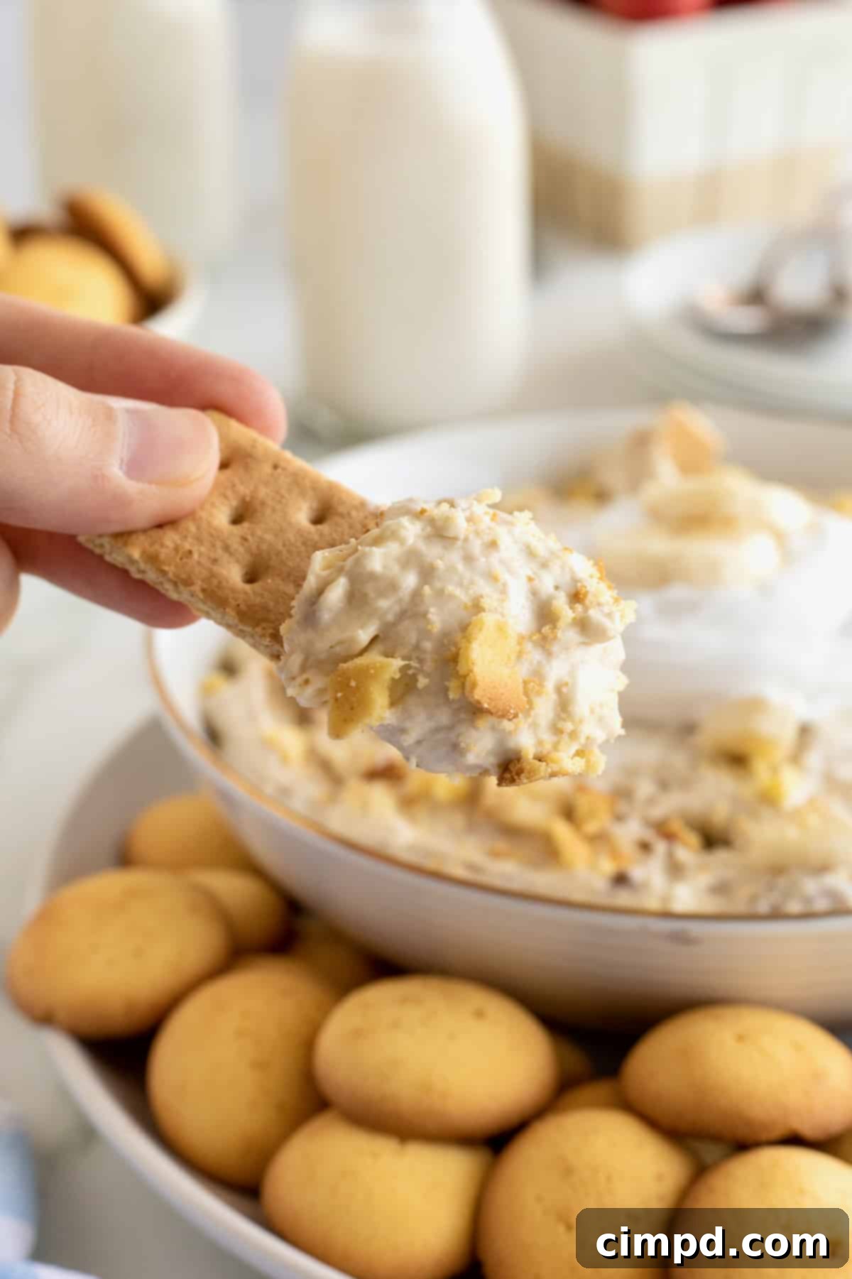 Dreamy Banana Pudding Dip 3 A hand dipping a graham cracker into a large white bowl of banana pudding dip.