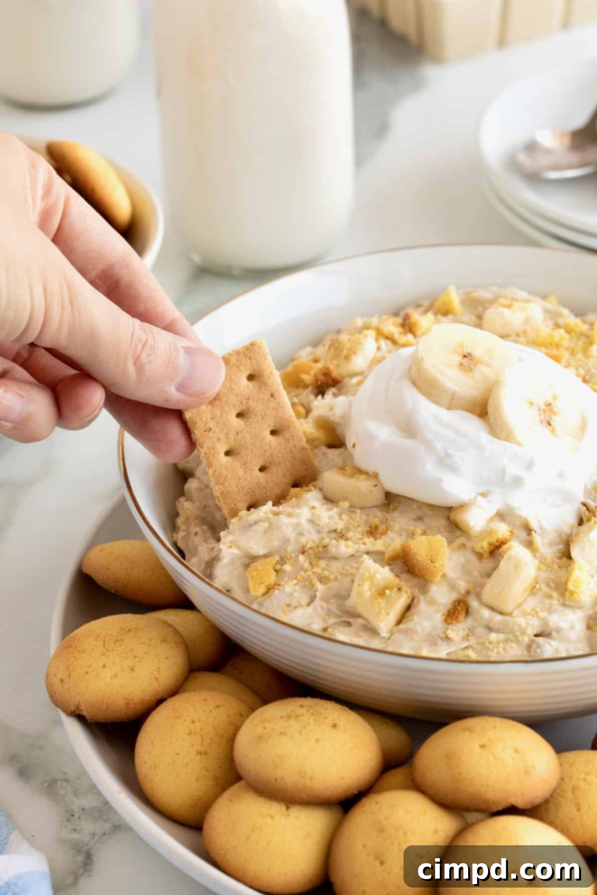 Dreamy Banana Pudding Dip 4 A hand dipping a graham cracker into a large white bowl of banana pudding dip.
