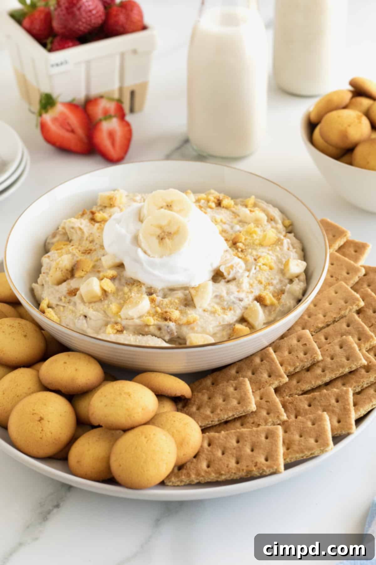 Dreamy Banana Pudding Dip 8 A bowl of banana pudding topped with whipped cream and two banana slices surrounded by graham crackers and Nilla wafers.