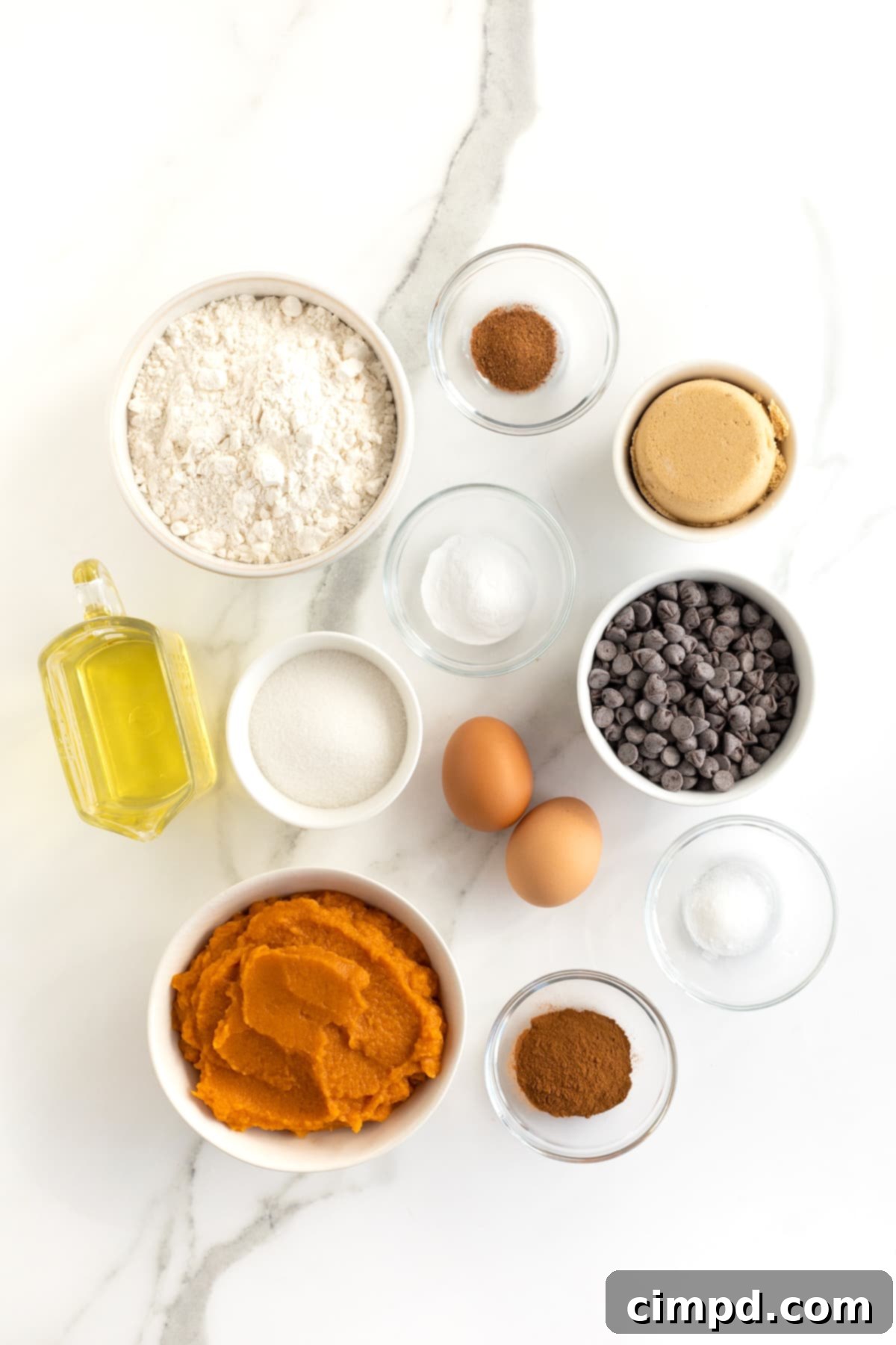 A collection of ingredients for pumpkin chocolate chip muffins, including flour, sugars, spices, oil, eggs, pumpkin puree, and chocolate chips, neatly arranged in small glass bowls.