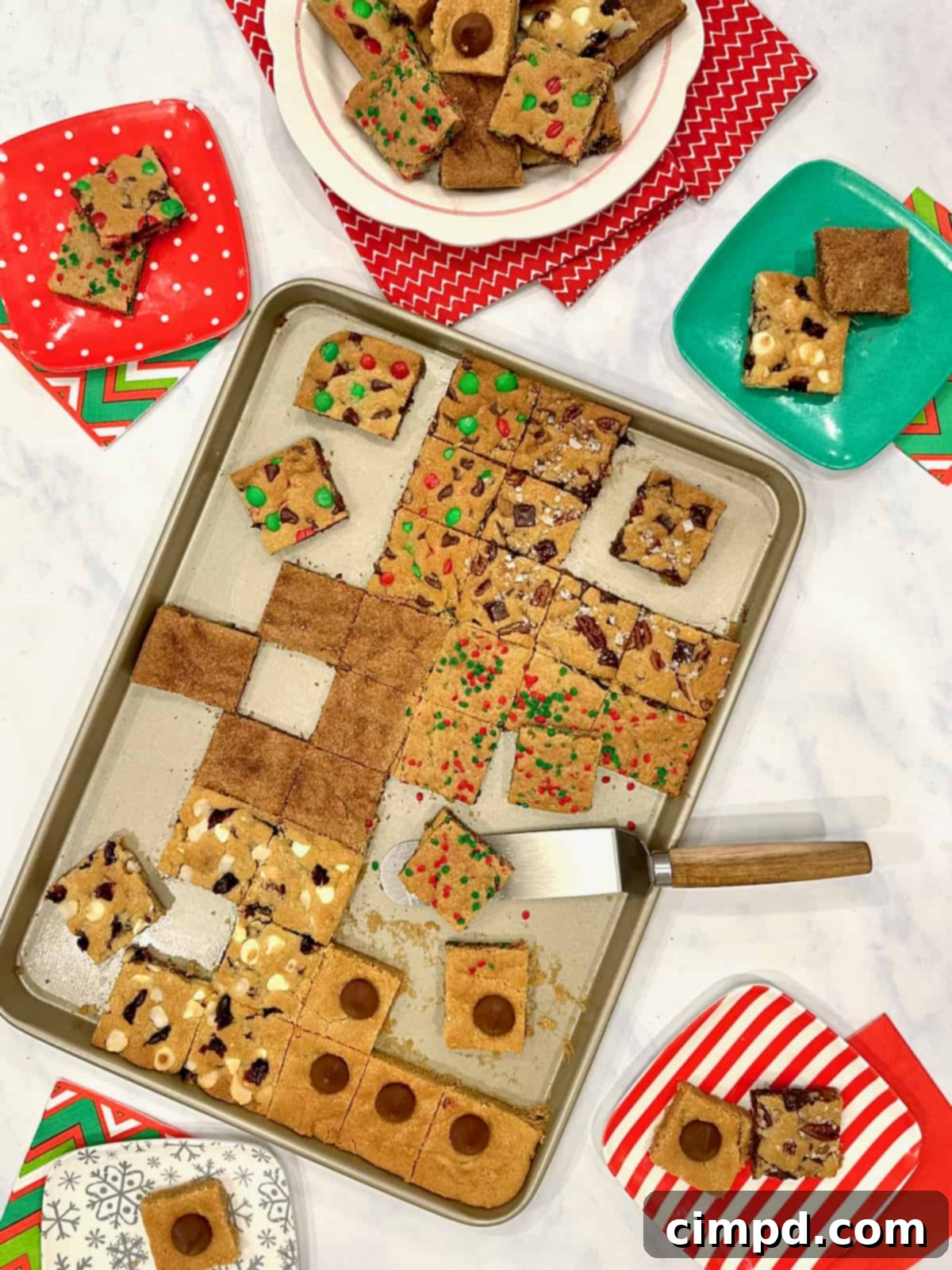 An aluminum baking sheet filled with an assortment of freshly baked holiday cookie bars, positioned beside a white plate showcasing more of the delicious treats.