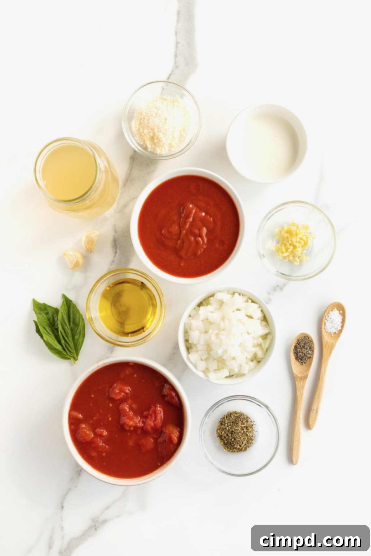 All the fresh ingredients for making homemade creamy tomato soup are neatly arranged in small white glass dishes on a pristine white counter top.