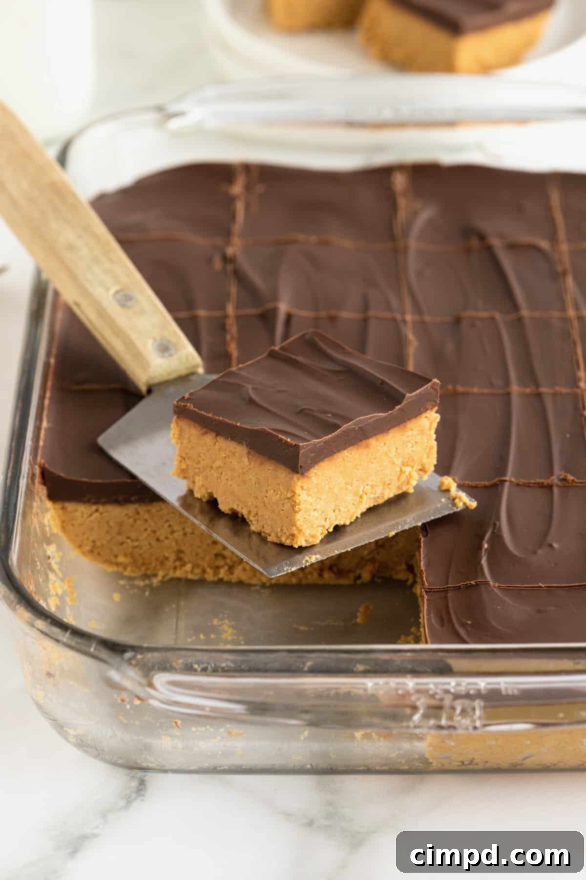 No Bake Peanut Butter Chocolate Bliss 3 A no-bake chocolate peanut butter bar on a metal spatula with a wooden handle in front of a baking dish of chocolate peanut butter bars.