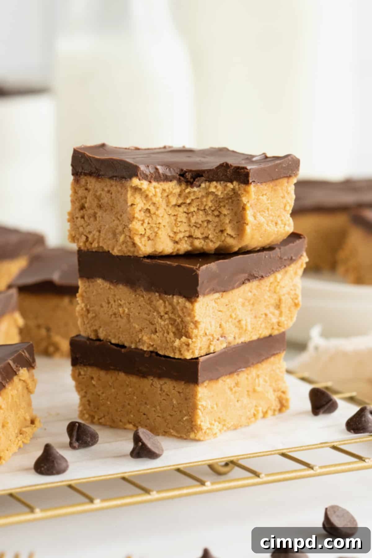 No Bake Peanut Butter Chocolate Bliss 4 Three chocolate peanut butter bars stacked on a parchment lined brass cooling rack with chocolate chips scattered around them. The top bar has a bite out of it.
