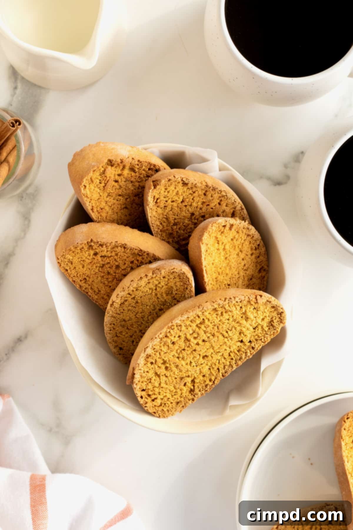 Six perfectly sliced pumpkin spice biscotti arranged in a white oblong serving dish, next to a steaming cup of coffee on a pristine white countertop.