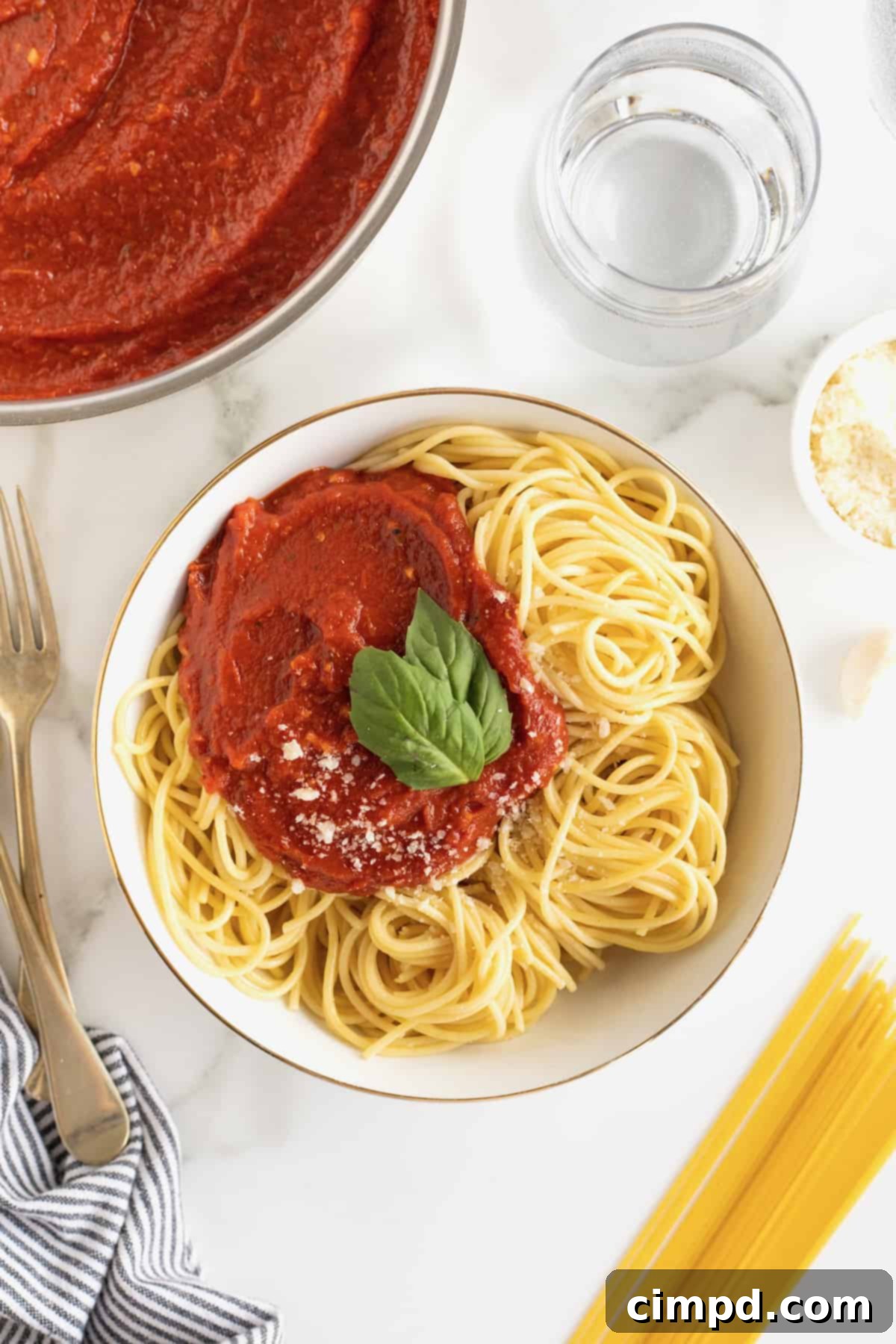 A beautifully plated bowl of cooked spaghetti, generously topped with vibrant red marinara sauce, fresh basil leaves, and a sprinkle of Parmesan cheese. A simmering pot of sauce is visible in the background.