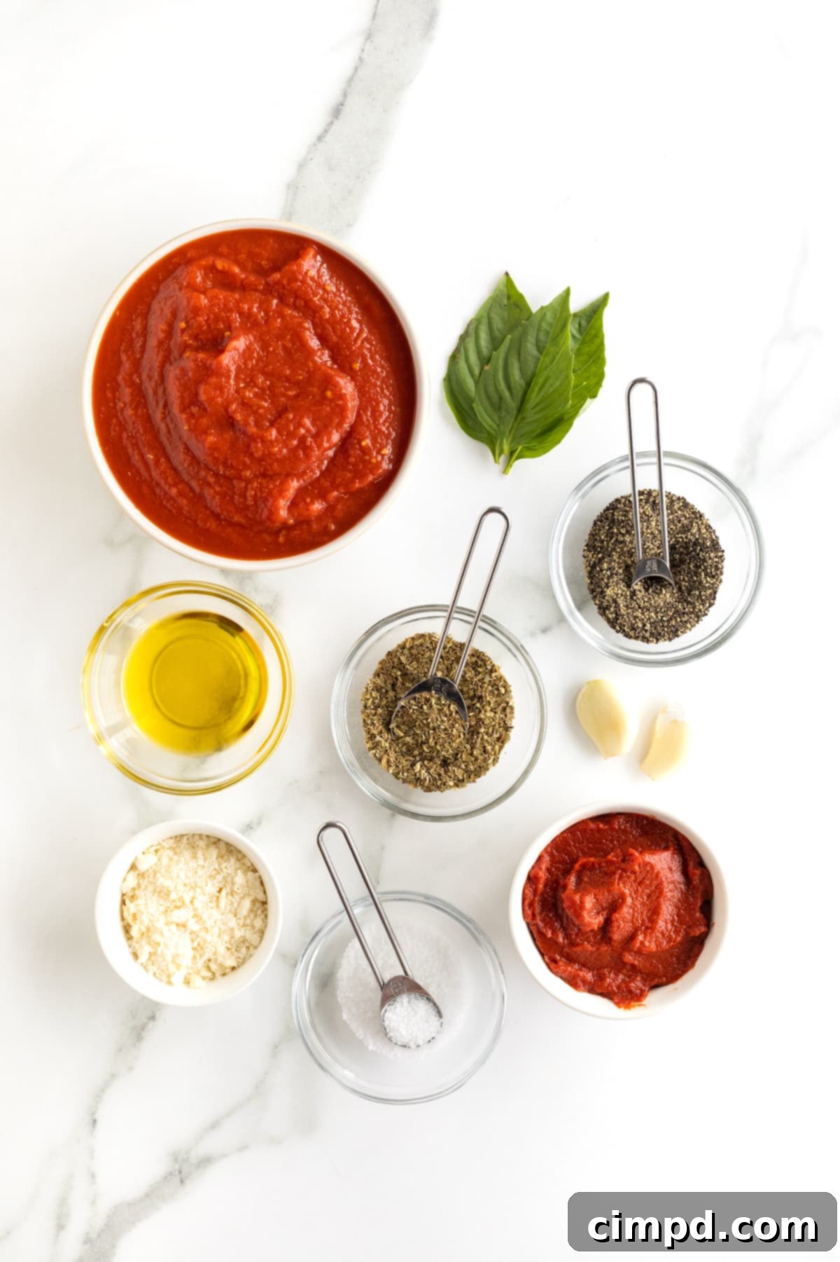 All the essential ingredients for simple marinara sauce artfully arranged in small glass dishes on a pristine white marble counter, with fresh basil and peeled garlic cloves directly on the surface.