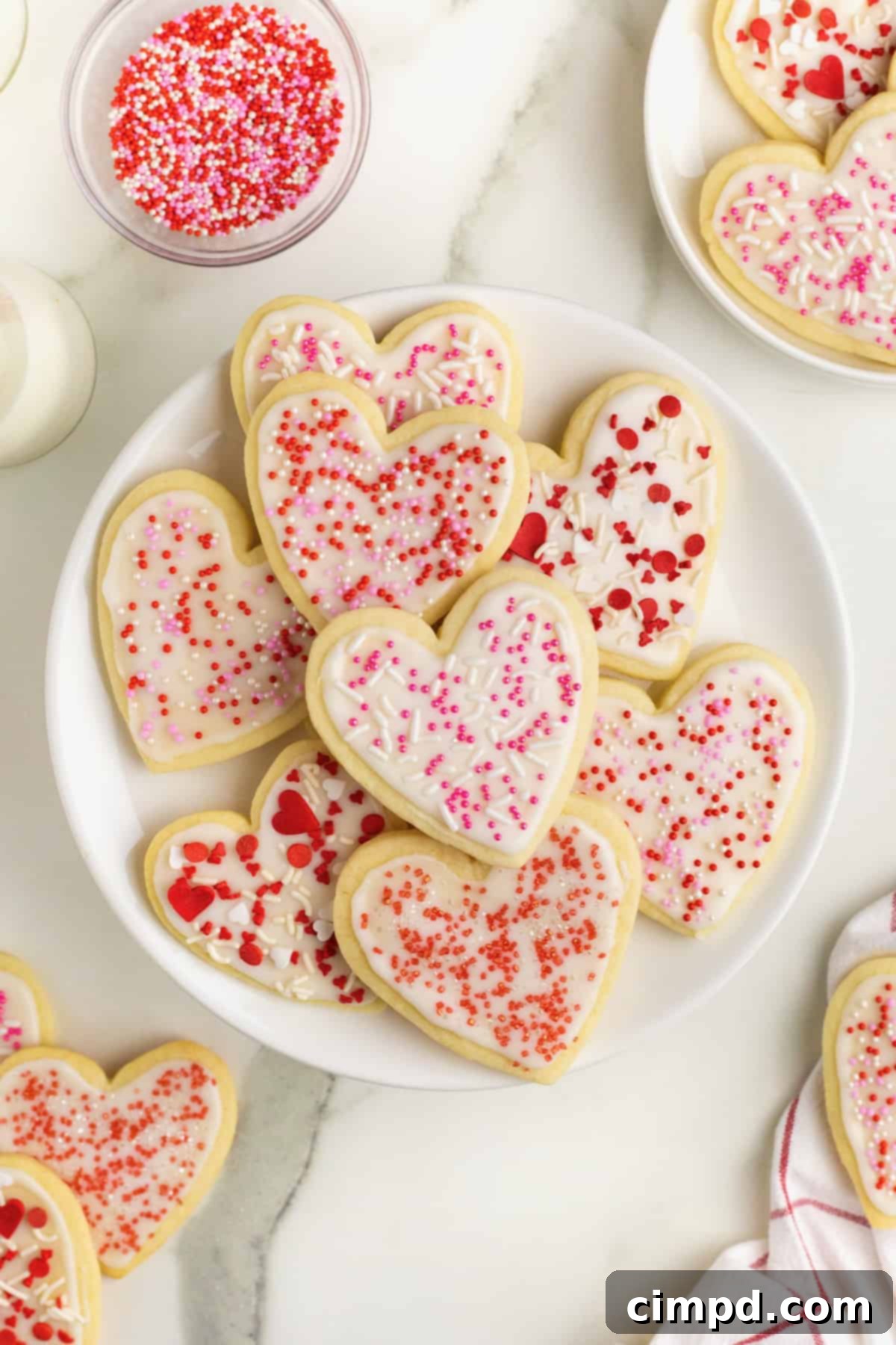 A charming pile of freshly baked heart-shaped sugar cookies, adorned with creamy white frosting and vibrant pink and red sprinkles, elegantly arranged on a large white serving plate.