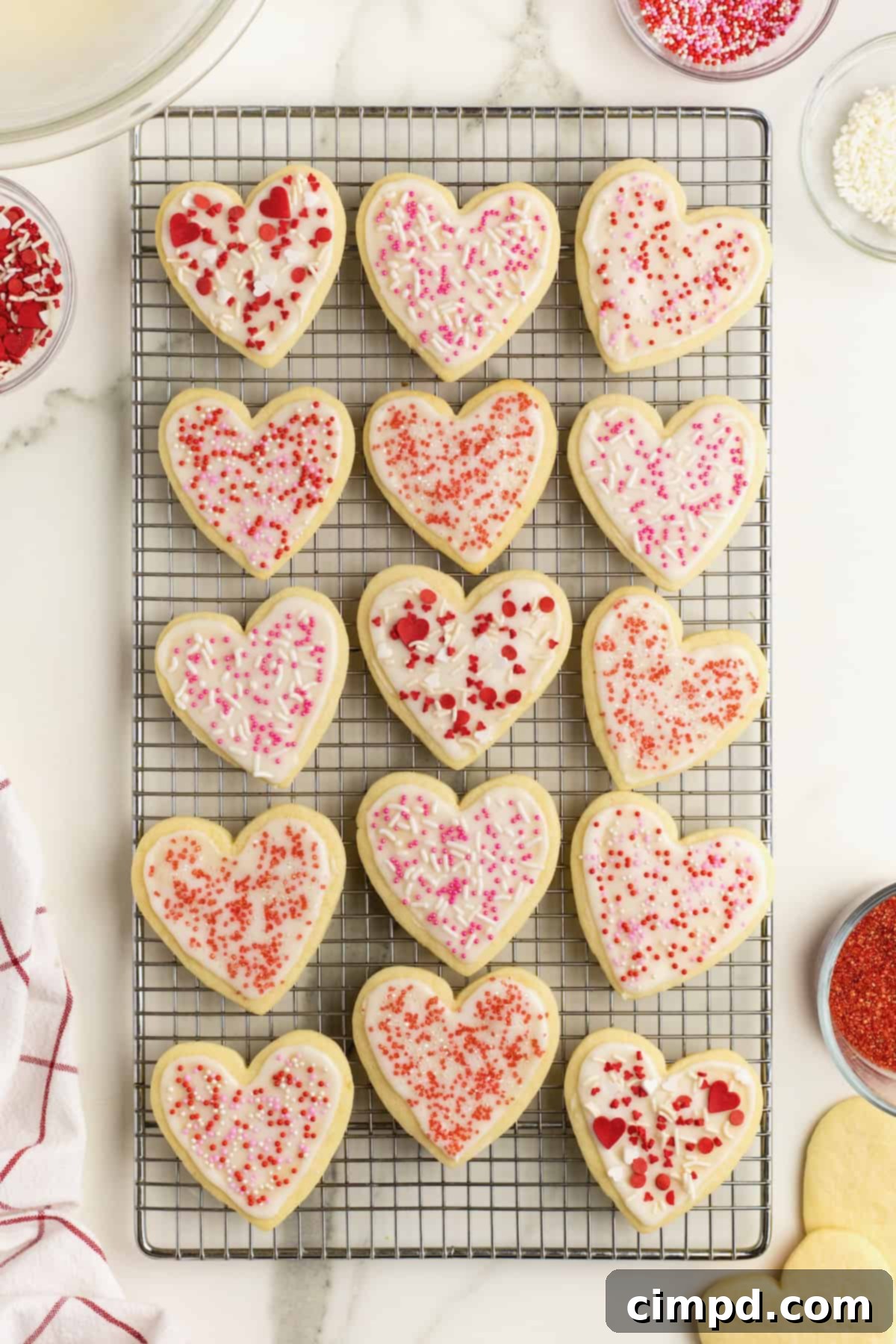 A delightful array of fifteen heart-shaped sugar cookies, each meticulously covered with pristine white frosting and adorned with a charming mix of red and pink sprinkles, neatly arranged on a wire cooling rack atop a sophisticated white marble counter, ready for enjoyment.