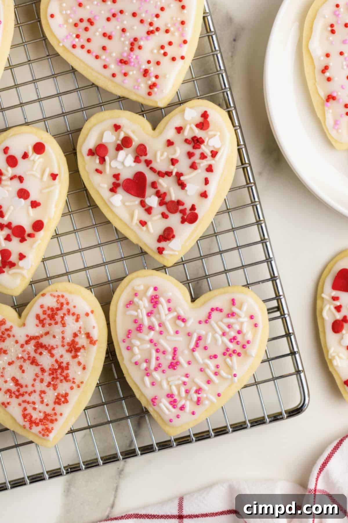 A wire cooling rack holds an inviting collection of heart-shaped sugar cookies, elegantly frosted with white icing and adorned with delightful red and pink sprinkles, all positioned on a pristine white marble counter, showcasing their readiness for enjoyment.