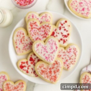 A pile of heart shaped sugar cookies with white frosting and pink and red sprinkles on a large white serving plate, optimized for social media sharing.