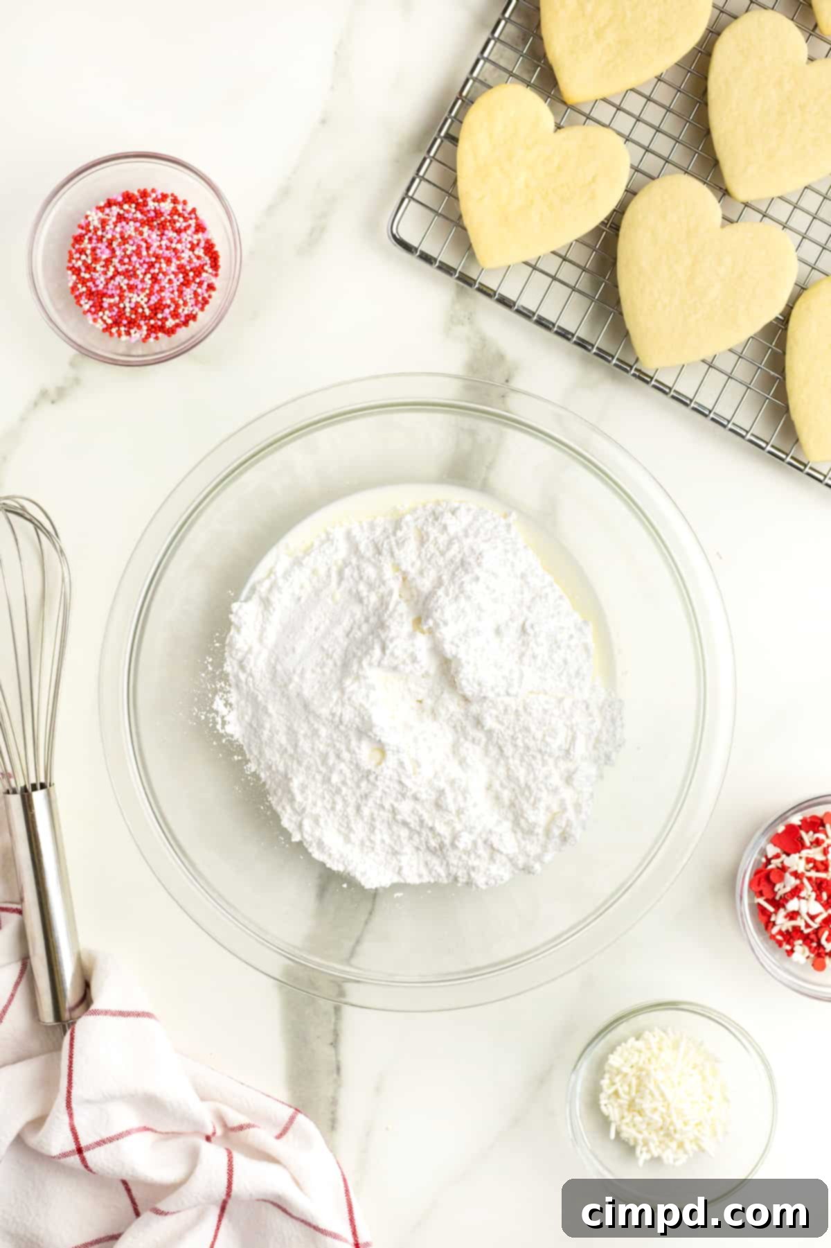 A clear glass bowl filled with the base ingredients for icing – powdered sugar and milk – positioned next to a wire cooling rack laden with heart-shaped sugar cookies. A wire whisk rests beside the bowl on a pristine white marble counter, illustrating the icing preparation stage.