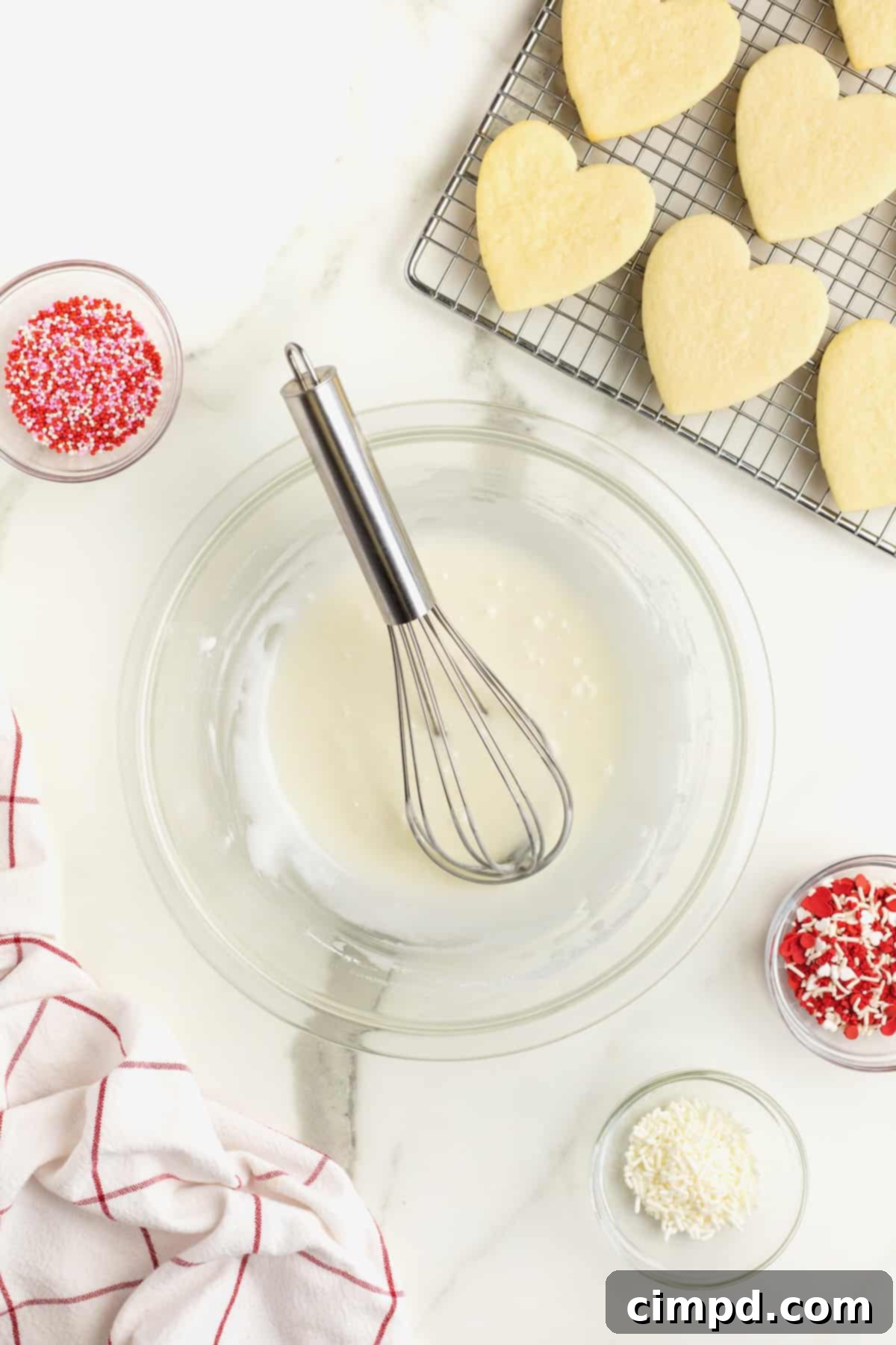 A glass bowl holds perfectly mixed icing, with a wire whisk inside, resting beside a wire cooling rack showcasing heart-shaped sugar cookies, all arranged on a elegant white marble counter, emphasizing the ideal icing consistency.
