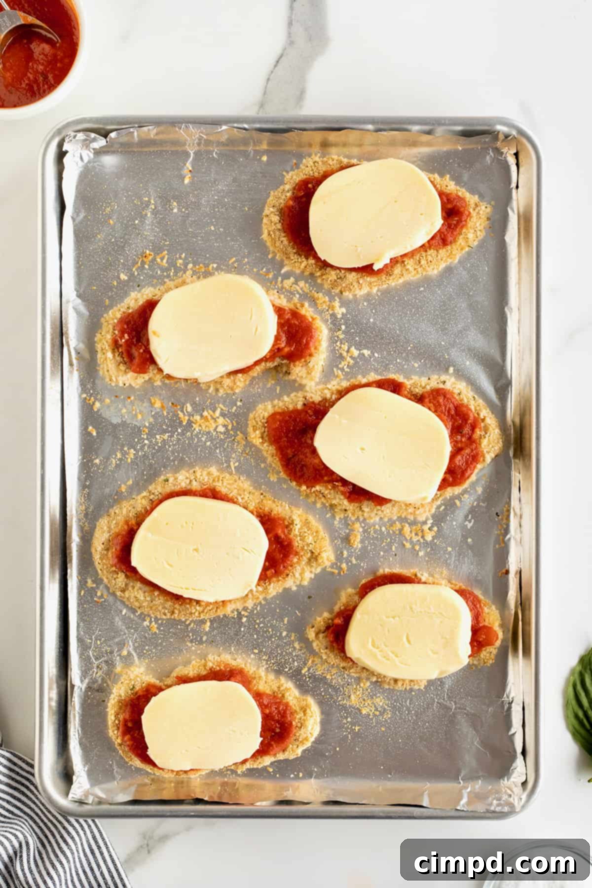 Six chicken cutlets topped with marinara sauce and fresh mozzarella on an aluminum baking sheet.