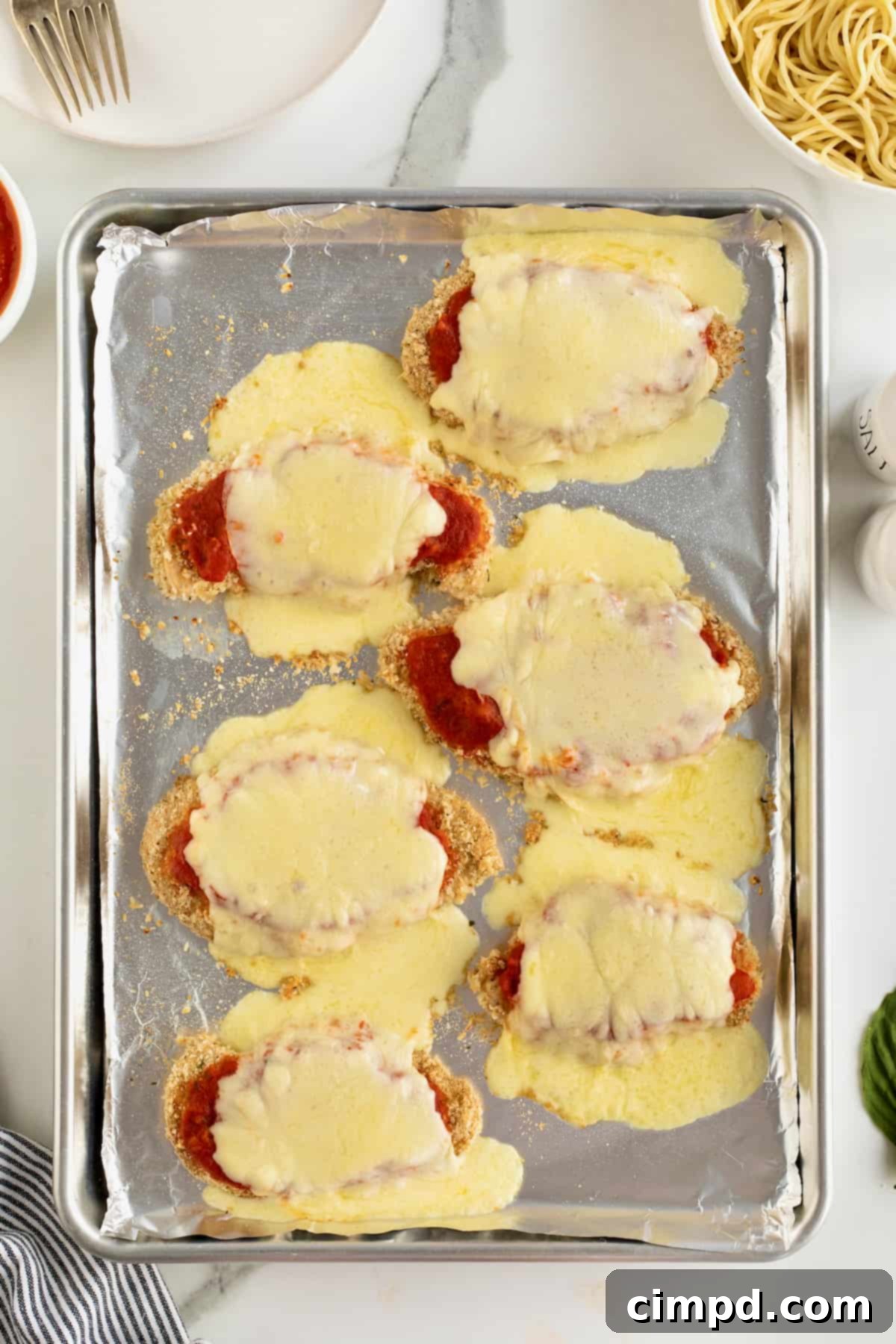 Six chicken cutlets topped with marinara sauce and melted fresh mozzarella on an aluminum baking sheet.