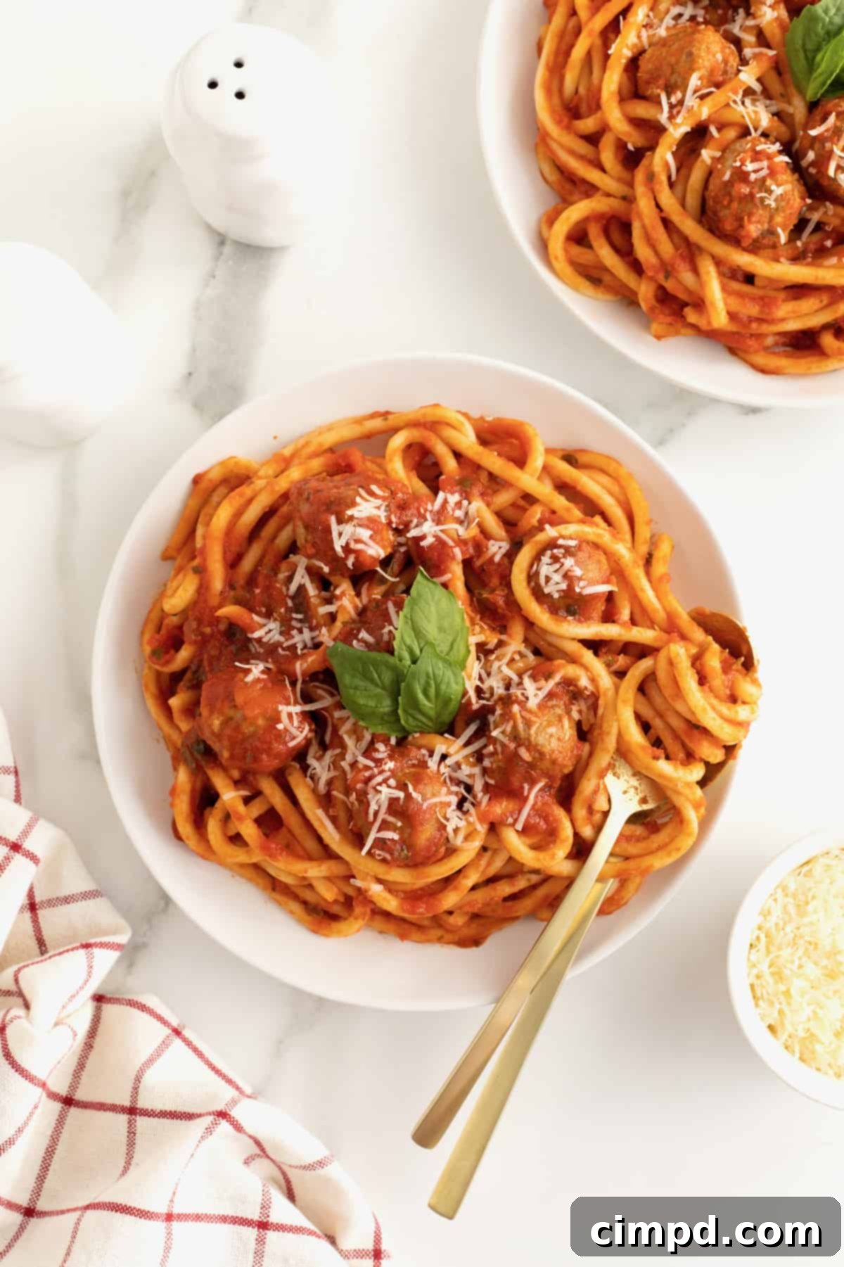 A large white serving bowl filled with spaghetti and meatballs. The spaghetti is garnished with three fresh basil leaves. 